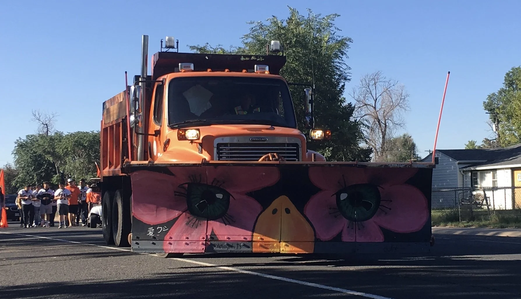 Orange snow plow truck with a front blade painted with pink flowers and green eyes, parked on a street. People are seen in the background.