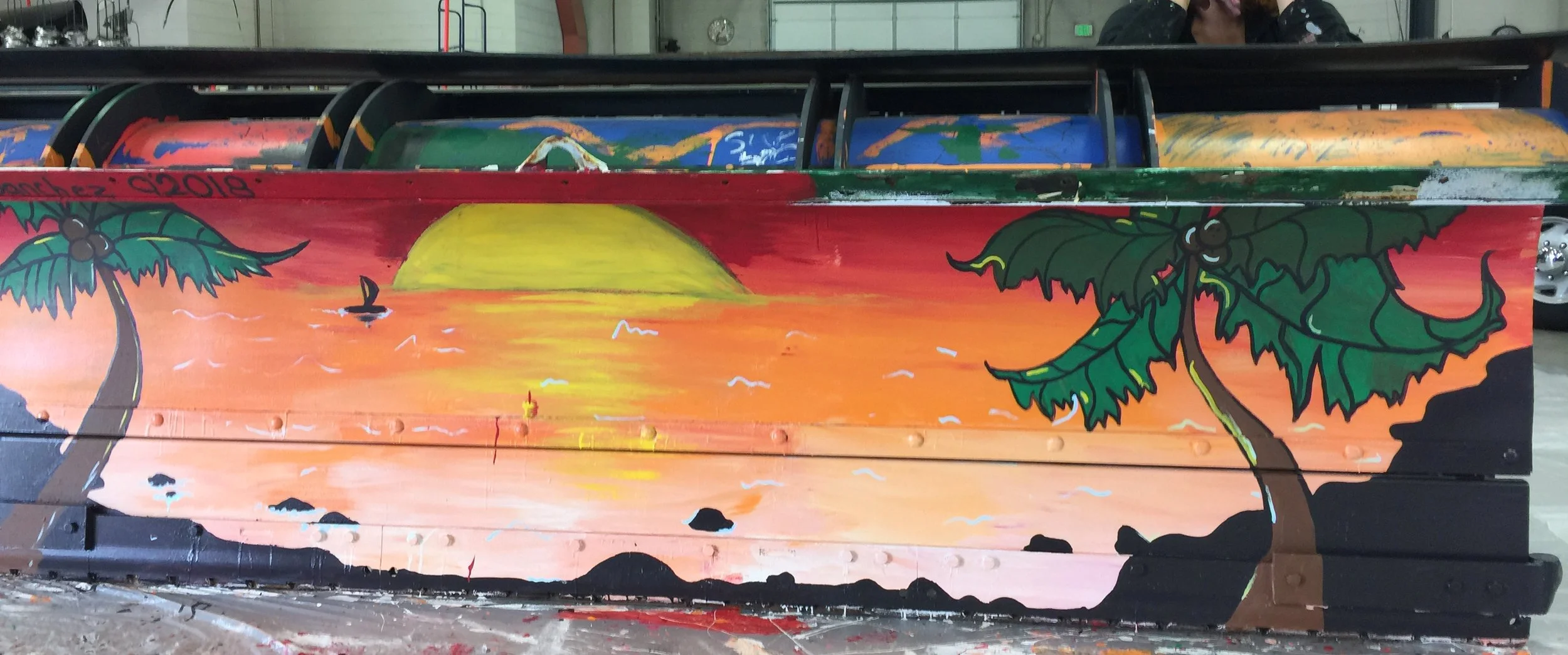 Colorful mural of a sunset with palm trees, birds, and ocean waves painted on a wooden surface.