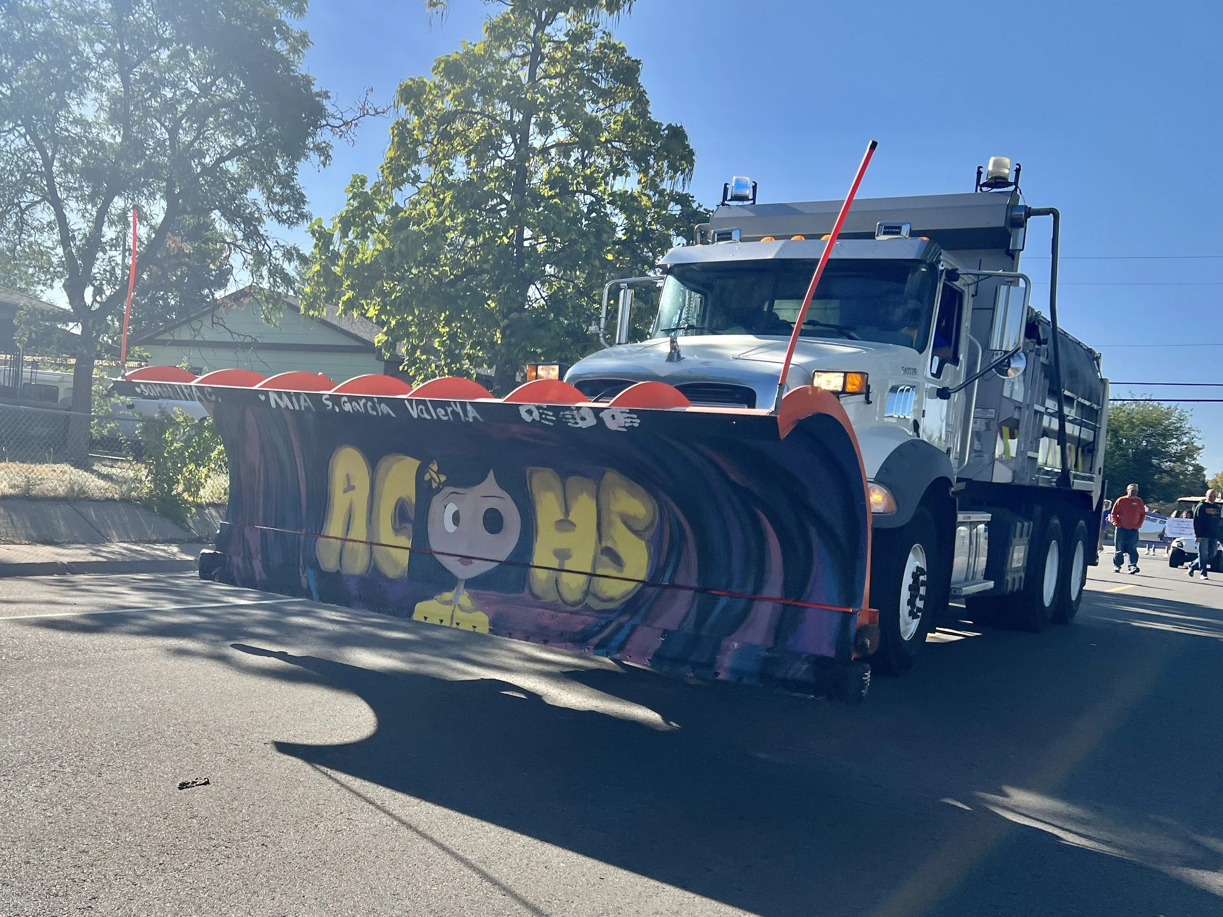 A street early in the morning with a large snowplow vehicle decorated with a colorful banner that says 'Ashe' and features a cartoon girl, with rock salt spreader blade in front of it, and people in the background.