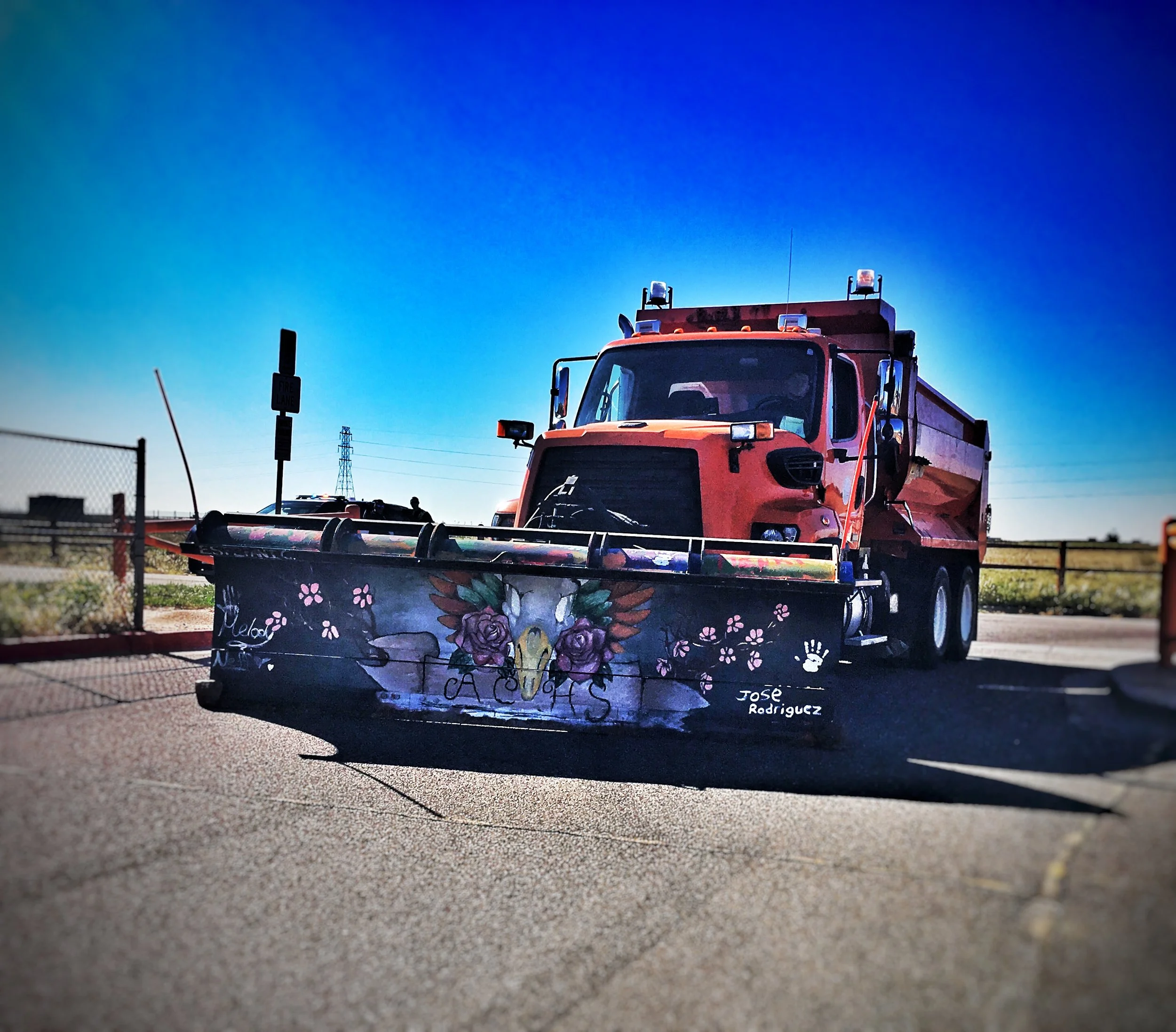 A large orange snowplow truck with a decorated plow featuring a colorful skull with flowers and the words "CA CUAS" parked on a paved lot under a clear blue sky.