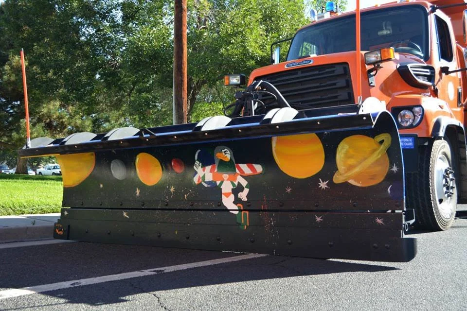 Orange snowplow truck with a space-themed design on the front blade, featuring planets, stars, and a cartoon astronaut bird.