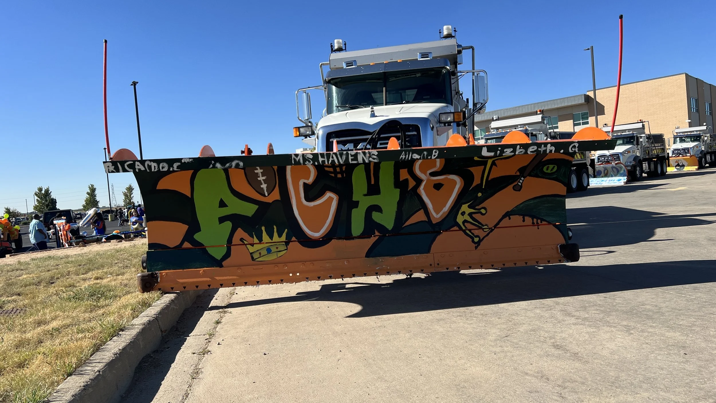 Large street sweeper truck with colorful graffiti artwork on the front blade, featuring the name 'Acho', a crown, and football symbols, parked outdoors on a sunny day with other trucks and people in the background.