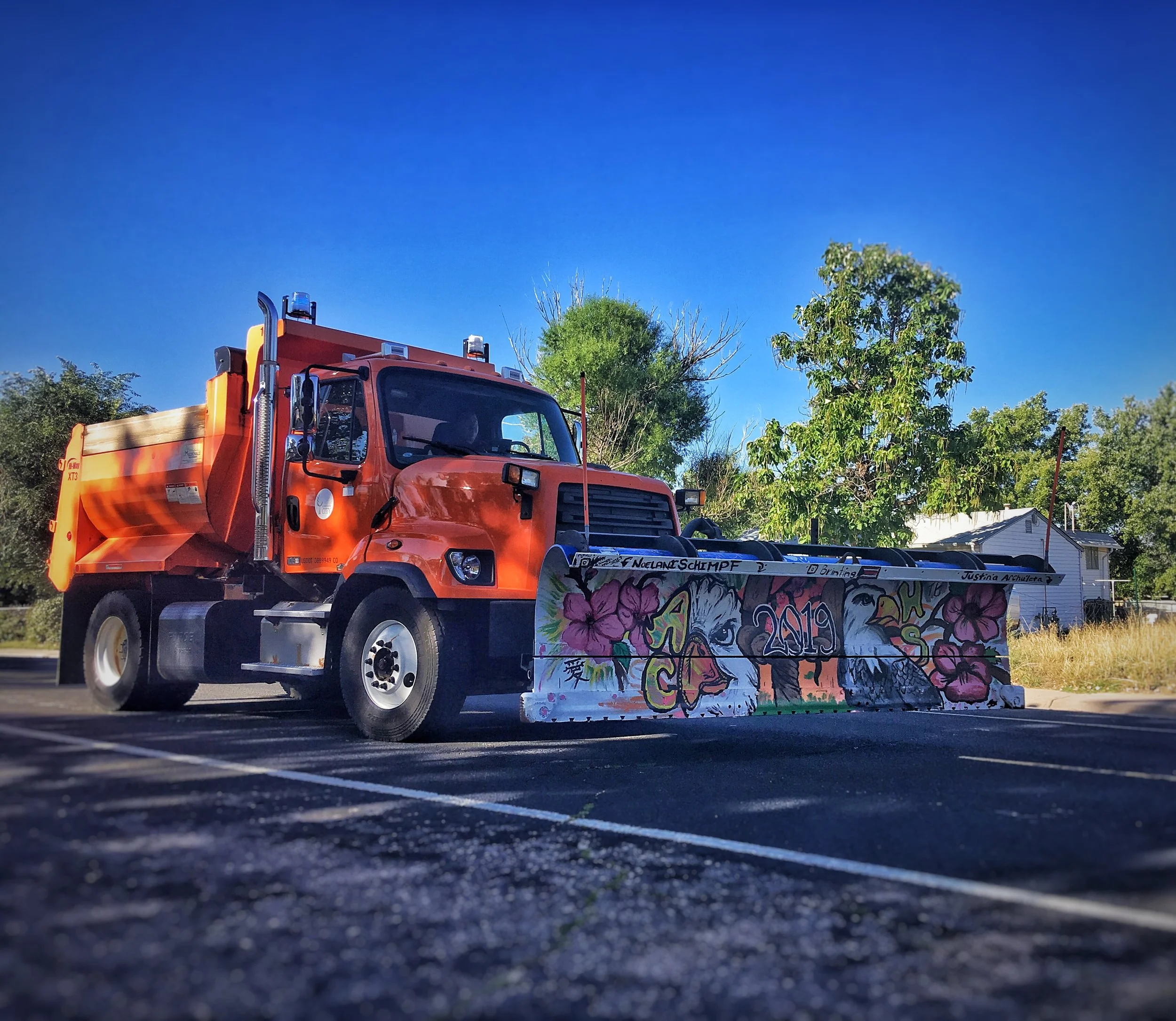 Orange snowplow truck with graffiti art on the front blade, parked on asphalt with trees, a house, and clear blue sky in the background.