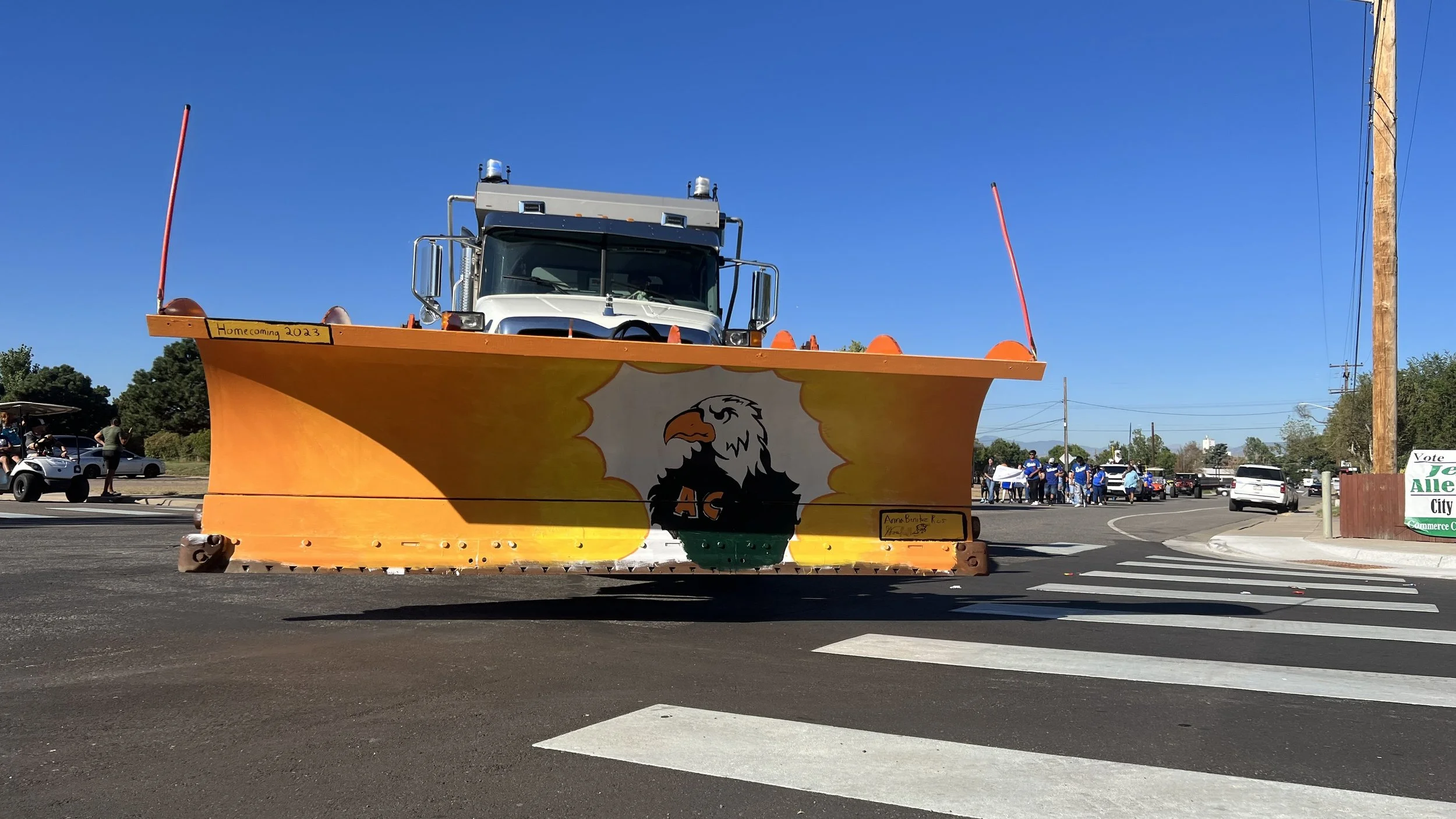 A large parade float designed to look like a boat with an orange hull and a white eagle logo with the letters 'AC' on its chest. The float has a truck on top and flags at each end. The float has small signs that read 'Homecoming 2023' and 'Anne Brigi