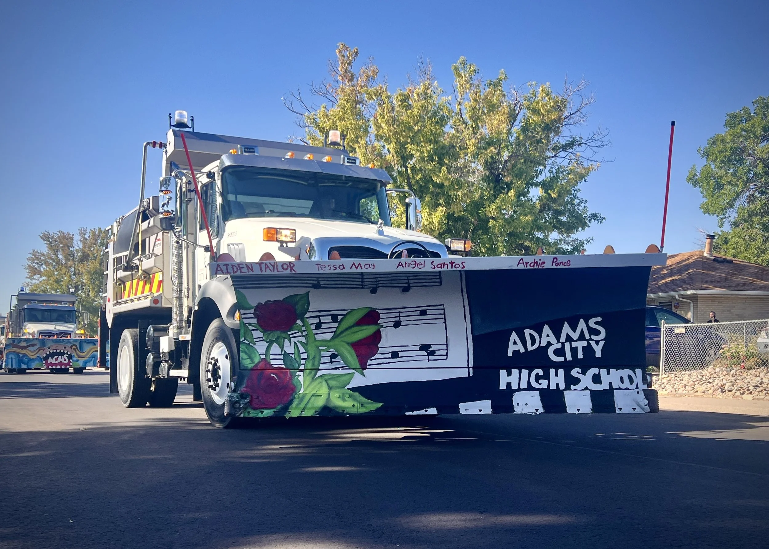 A parade float designed as a snow plow truck with a black and white front, decorated with red roses, green leaves, musical notes, and the words 'Adams City High School' on it. The float displays names of students written on top.