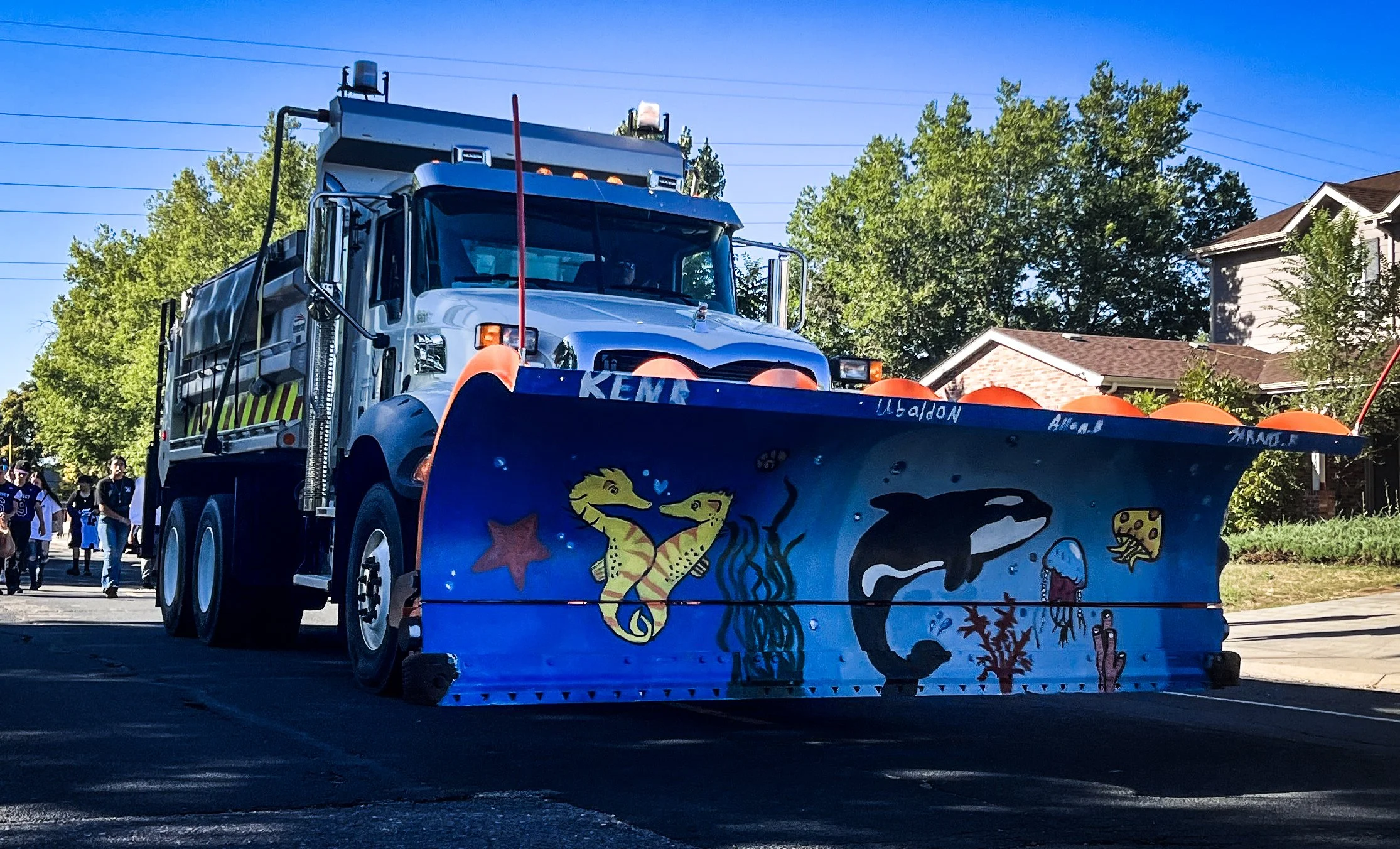 Fire truck with a colorful underwater scene painted on the front, including seahorses, a whale, algae, jellyfish, and coral, during a parade with people walking nearby and trees in the background.