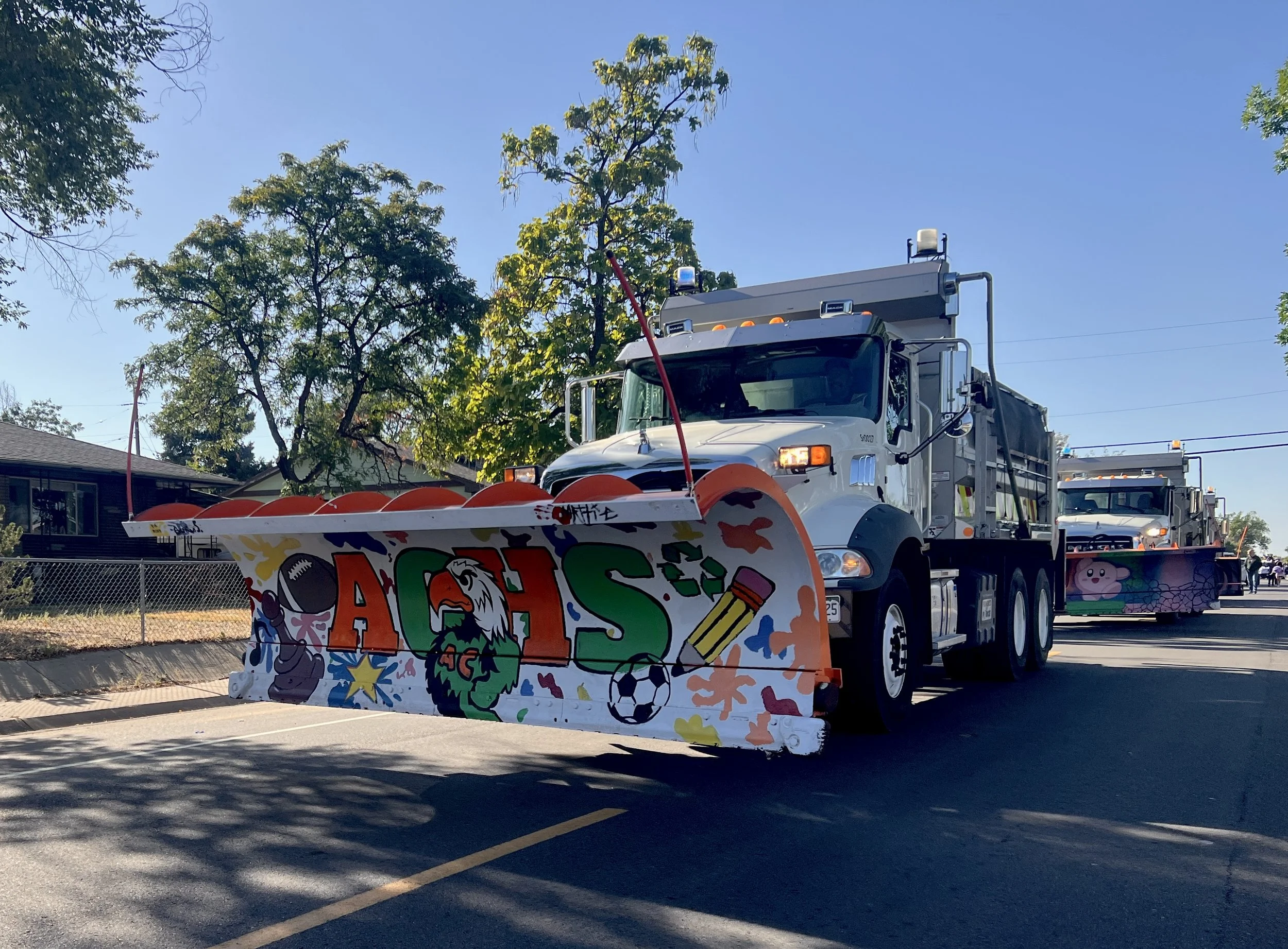A parade float decorated with a colorful, school-themed display featuring the letters 'ACHS' with a mascot, a football, and a soccer ball, driven by a large white fire truck with a snowplow attachment in front.