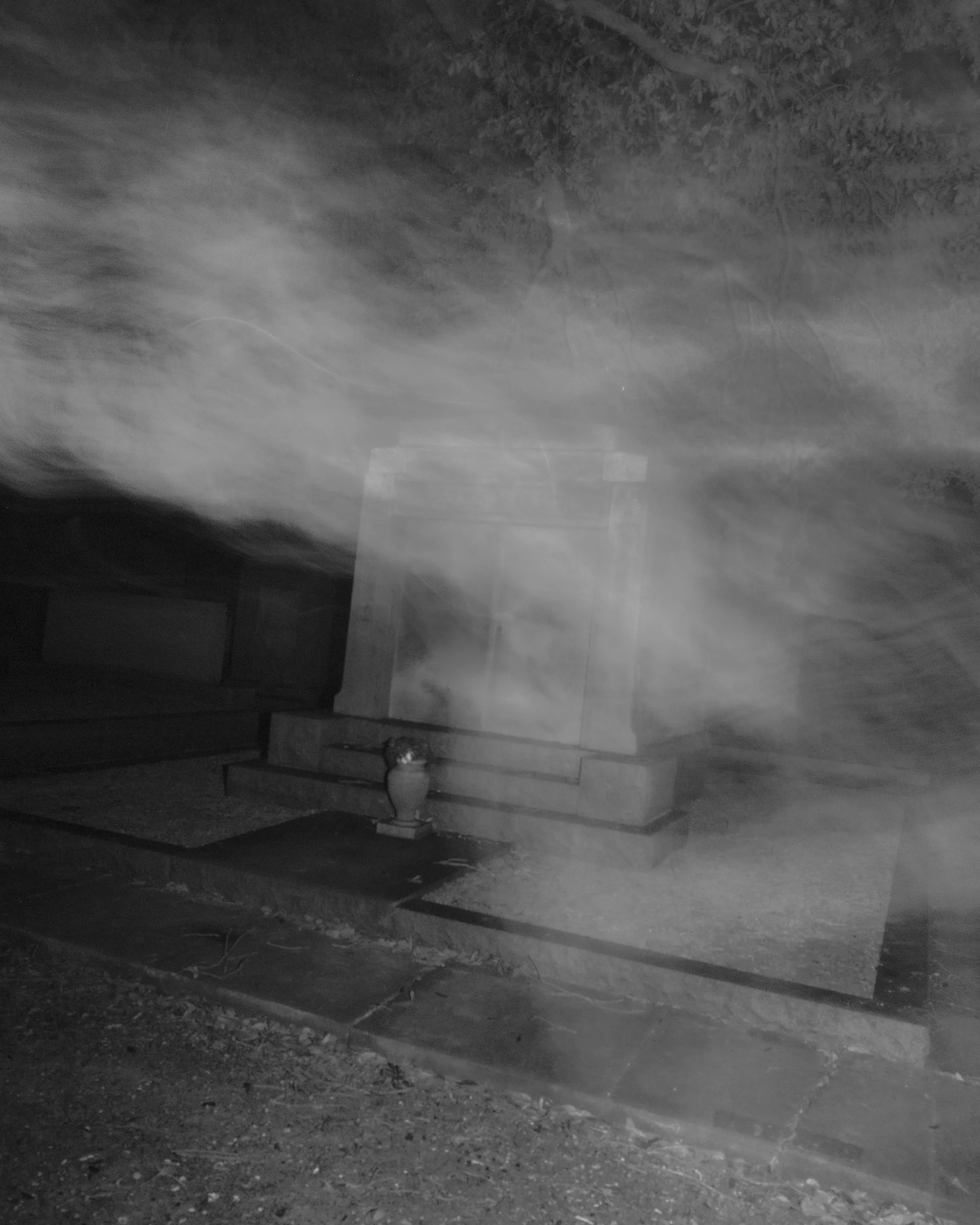 Nighttime image of a construction site or pathway with gravel, dirt, and a concrete curb. Visible in the background are trees, and there is a small white traffic cone or marker on the ground near the curb.