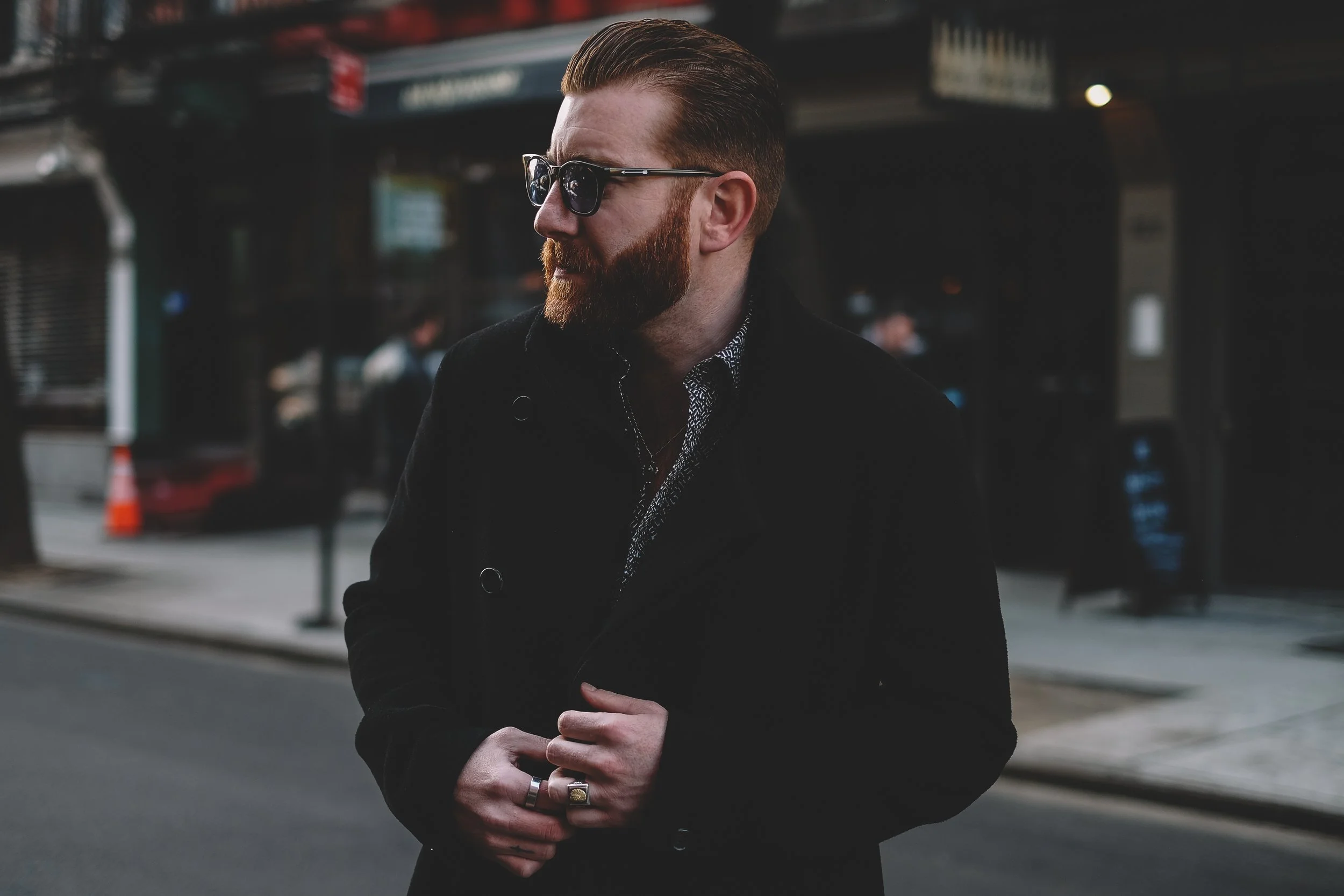 A man with a well-groomed beard, wearing sunglasses and a dark coat, standing on a city street during daytime.