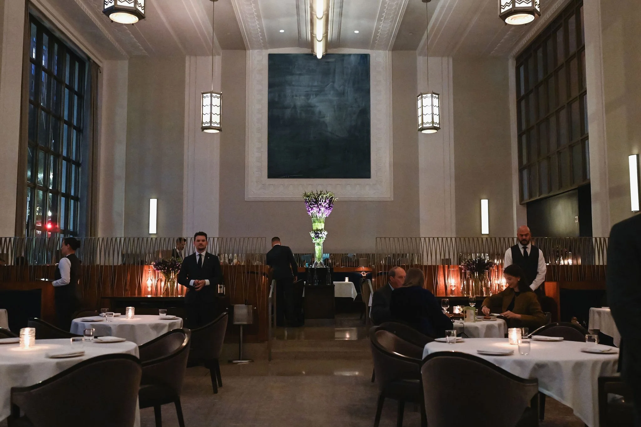 Restaurant dining area with tables set for dinner, waitstaff standing by, large abstract painting, tall windows, and elegant lighting.