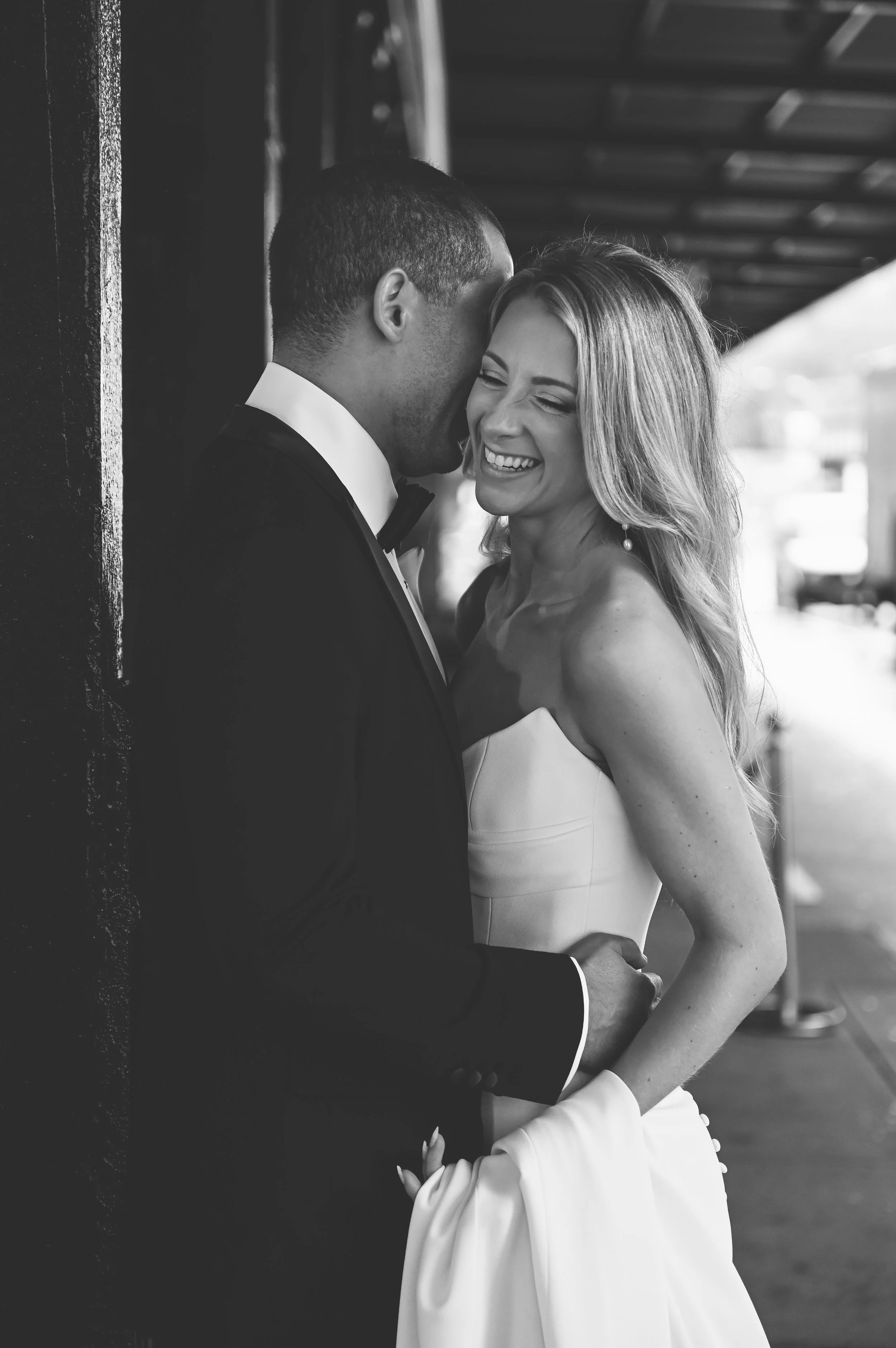 A black and white photo of a wedding couple, the groom wearing a tuxedo and the bride in a strapless wedding gown, smiling and sharing a tender moment outdoors.