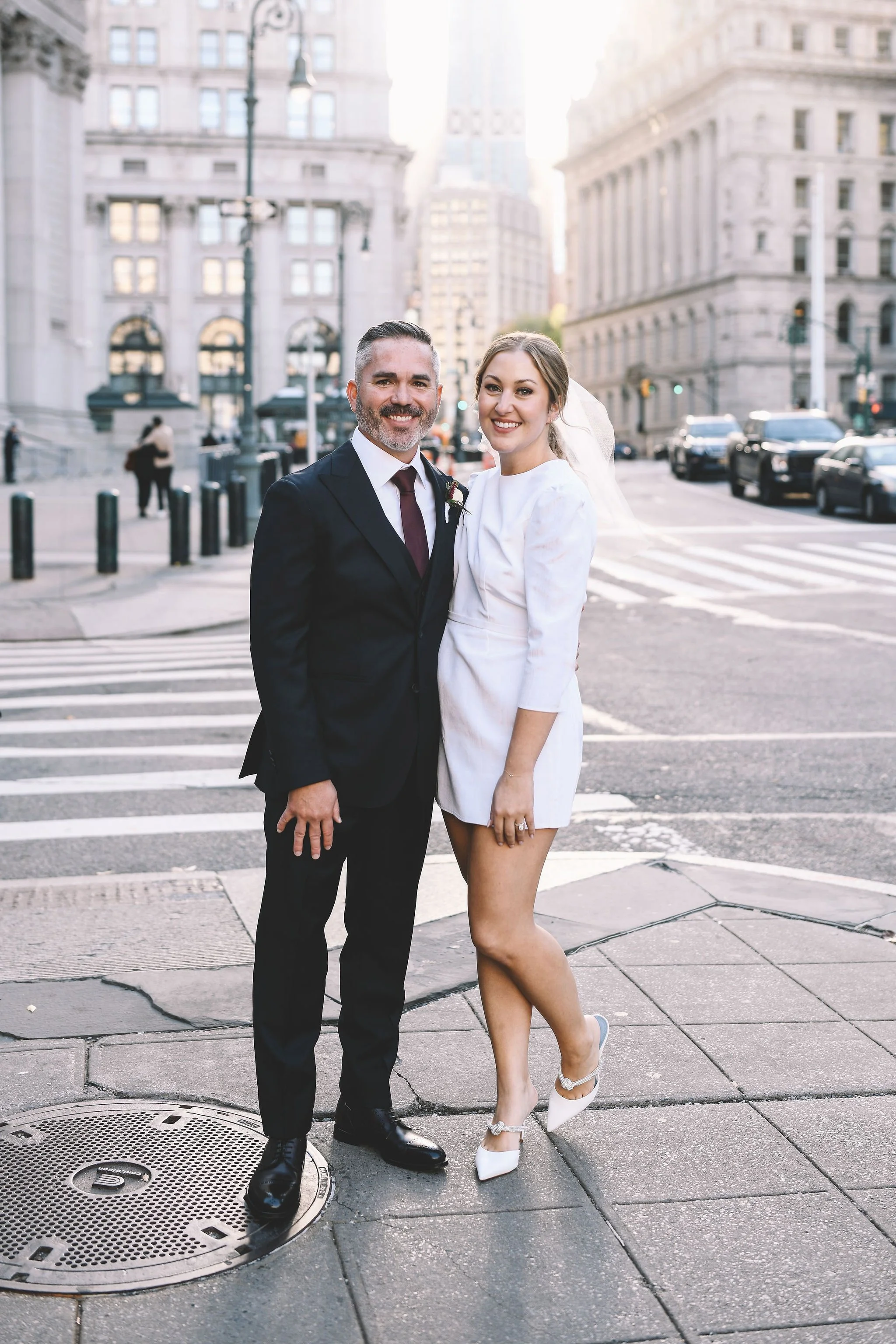 A man and woman in wedding attire stand on a city street corner, smiling at the camera with tall buildings and cars in the background.