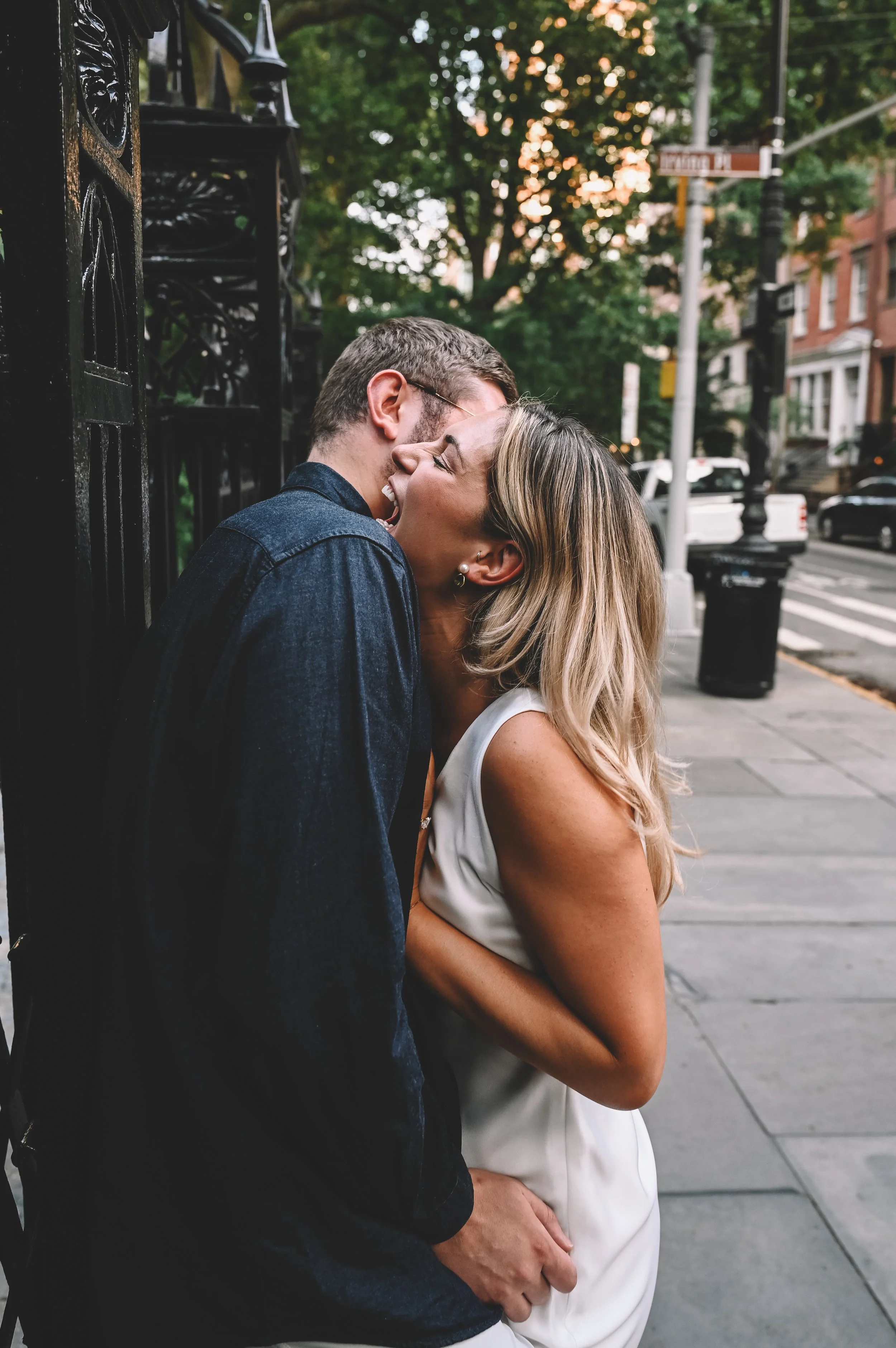 A couple sharing a passionate kiss on a city sidewalk at sunset, with trees and buildings in the background.