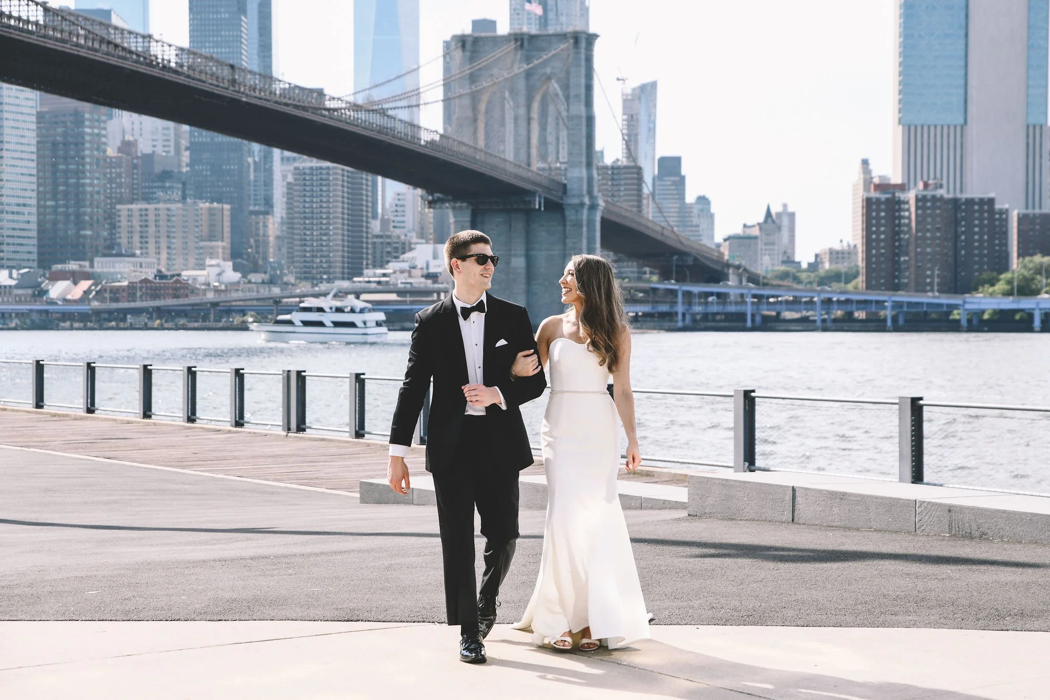 A newlywed couple walking along a waterfront with the Brooklyn Bridge and New York City skyline in the background. The groom is in a tuxedo and the bride in a white gown.