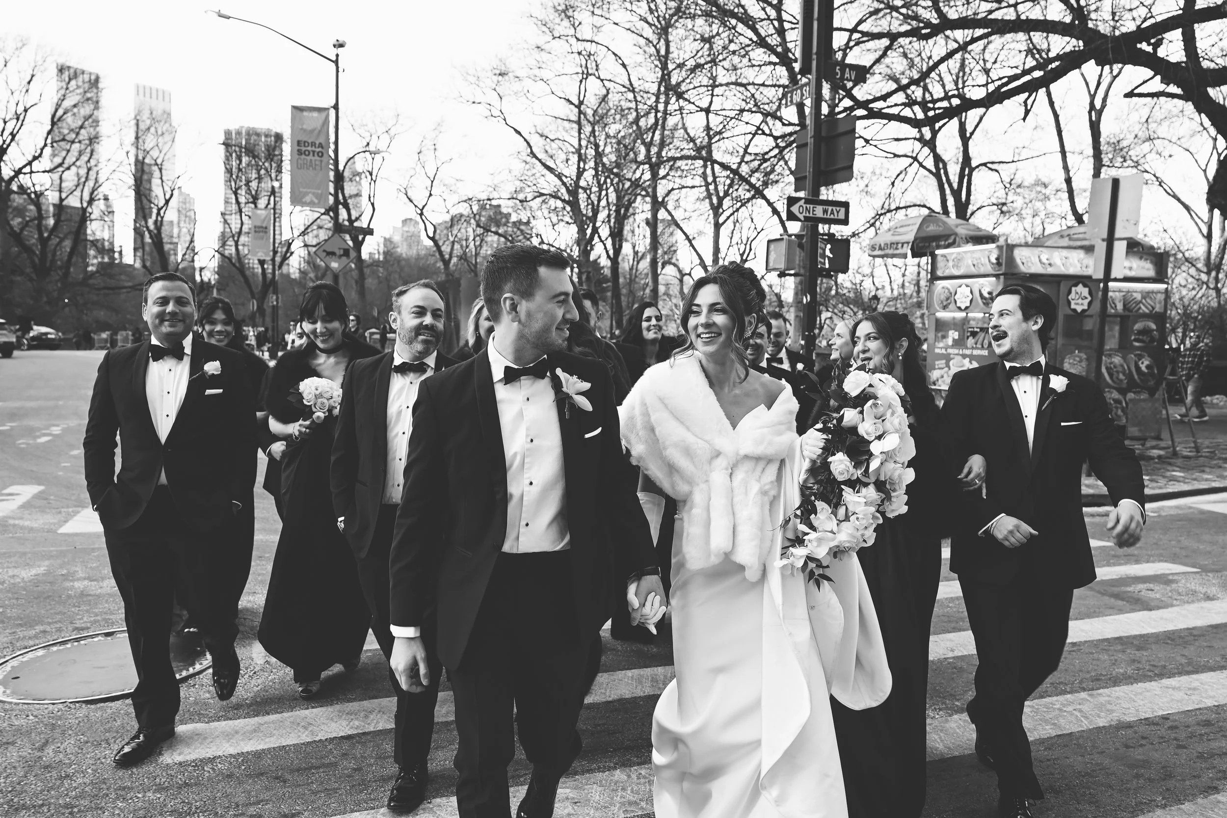 Black and white photo of a bride and groom walking on a city street, surrounded by friends, all dressed in formal wedding attire, smiling and holding bouquets.