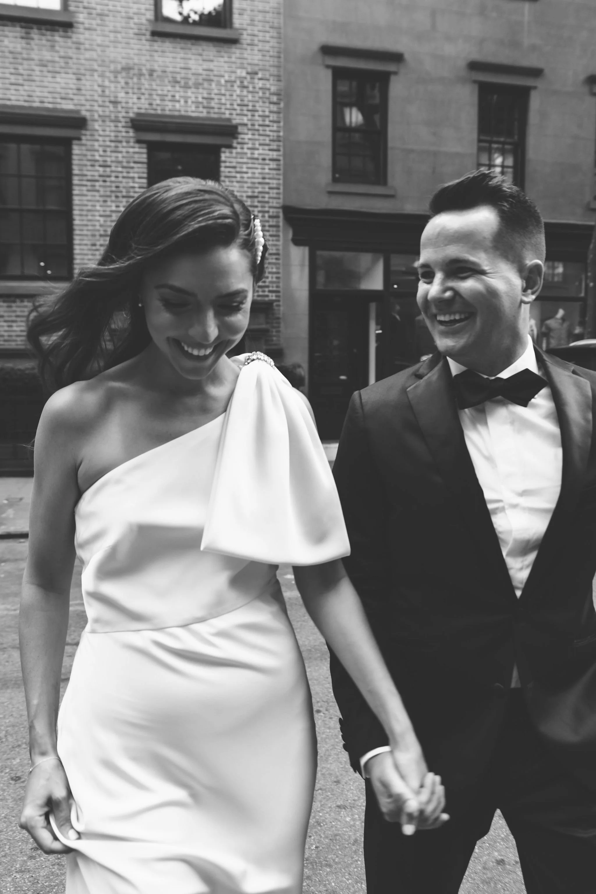 A smiling bride and groom holding hands walk outdoors in black and white, with brick and building windows in the background.