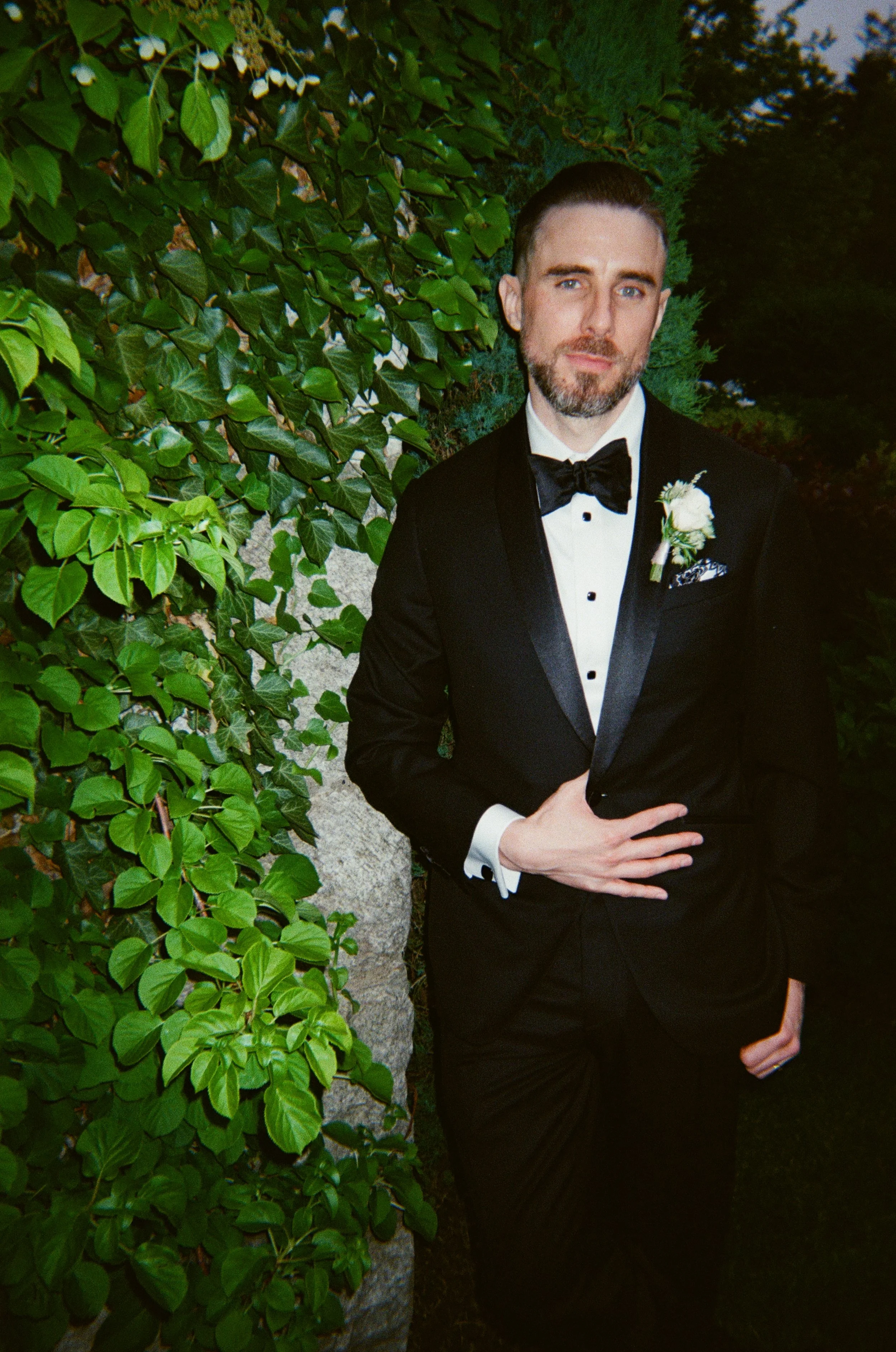 A man in a black tuxedo with a white shirt and black bow tie, standing outdoors against green foliage, with a boutonnière on his left lapel.