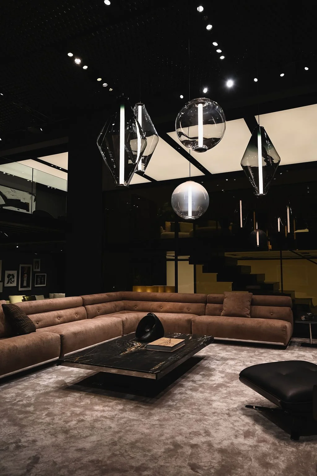 Modern living room with a large velvet brown sectional sofa, a black marble coffee table, and unique hanging glass pendant lights. The space features dark walls, artwork, and a plush area rug.