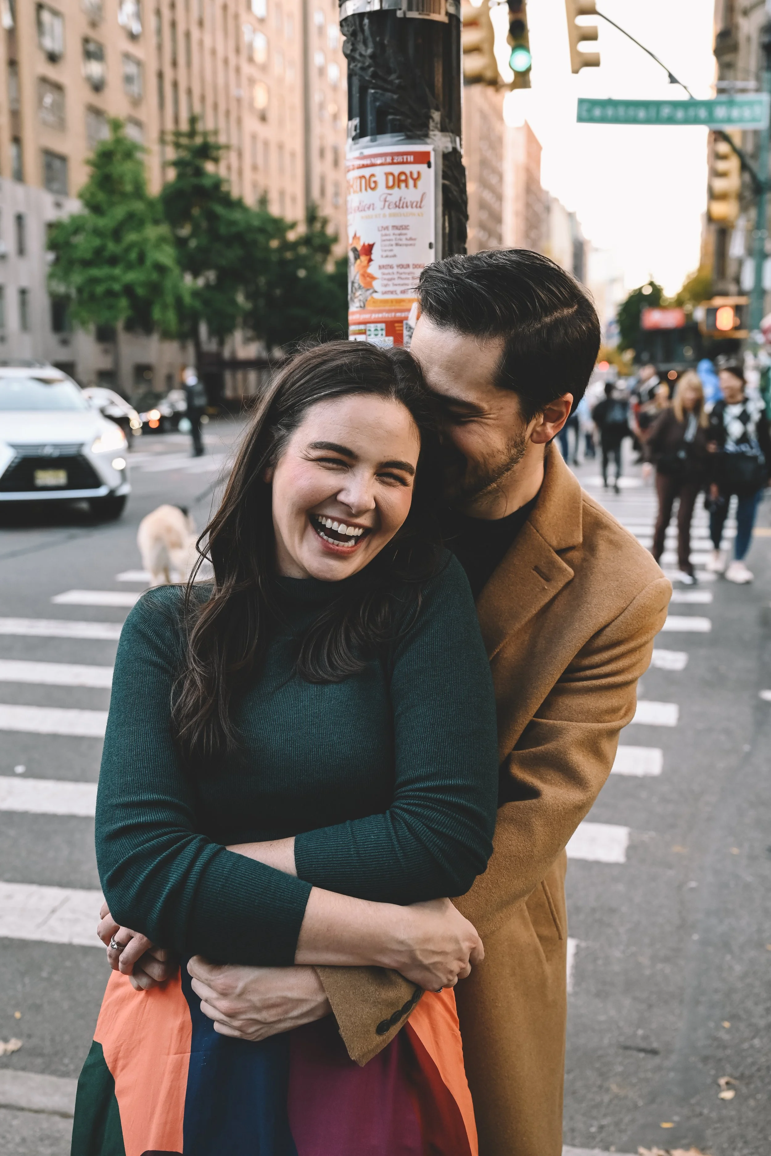 A happy couple hugging and smiling in a city crosswalk, with cars, pedestrians, and buildings in the background.
