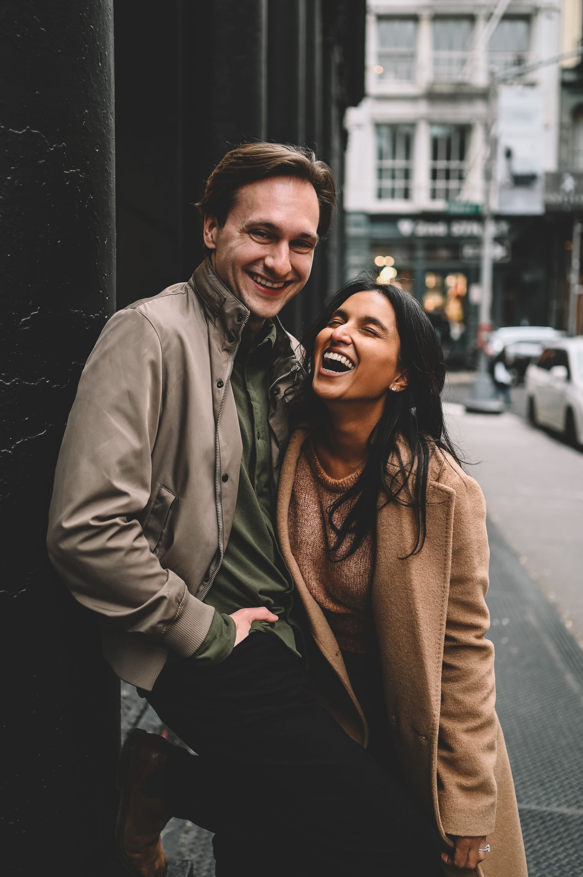 A smiling man and woman laughing together leaning against a wall on a city street.