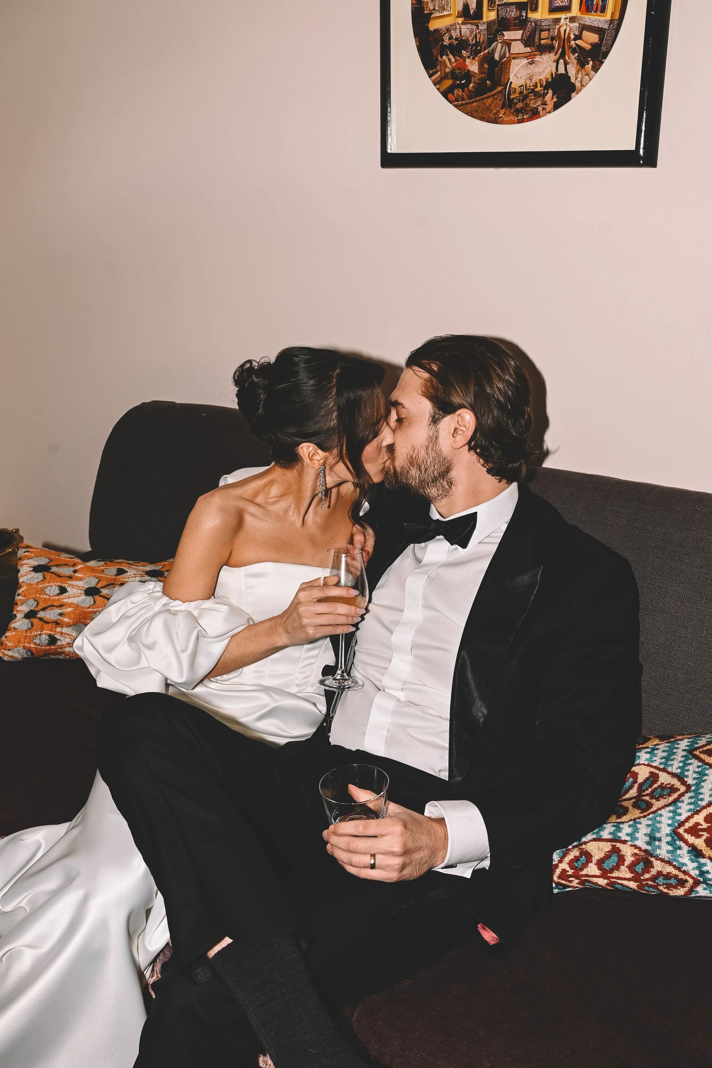 A romantic couple dressed in formal attire sharing a kiss while sitting on a dark-colored sofa. The woman is wearing a white gown, and the man is in a black tuxedo. They are holding champagne glasses.