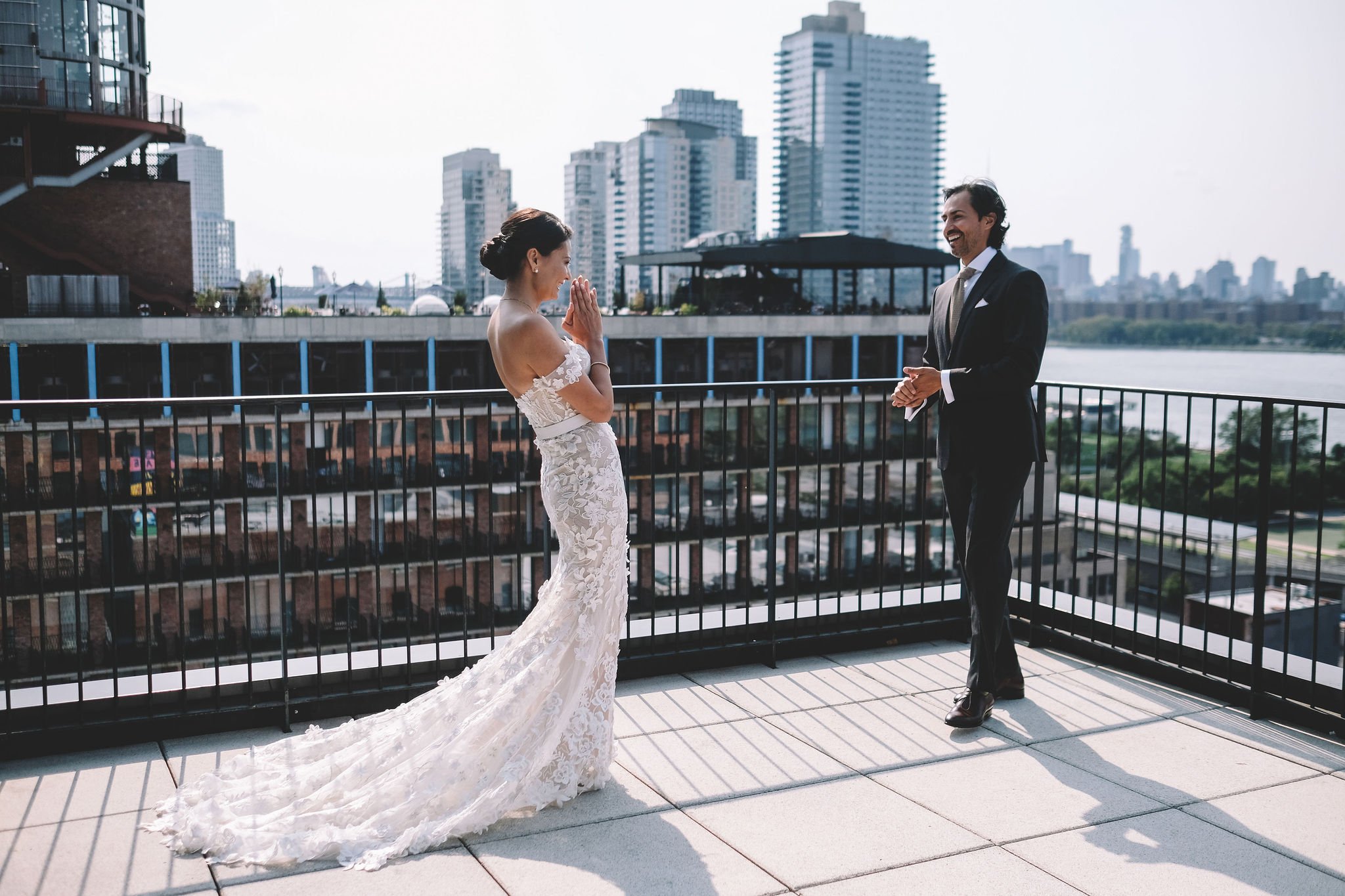 A bride and groom on a rooftop balcony, with a city skyline in the background. The bride is in a white lace wedding gown with a train, and the groom is dressed in a black suit with a white shirt and tie, smiling and facing each other.