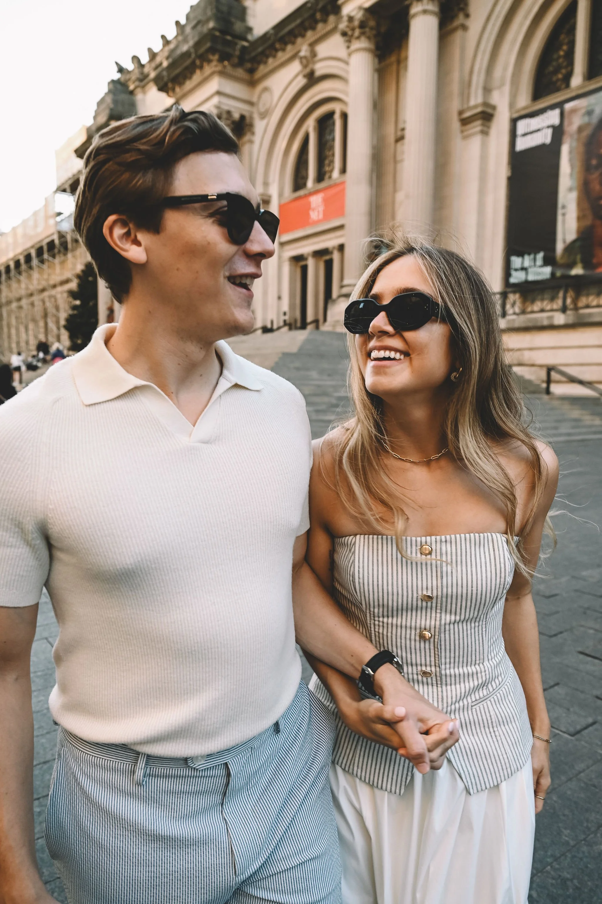 A young couple wearing sunglasses walking together, with the man holding the woman's hand, in front of a historic building with stairs and columns.