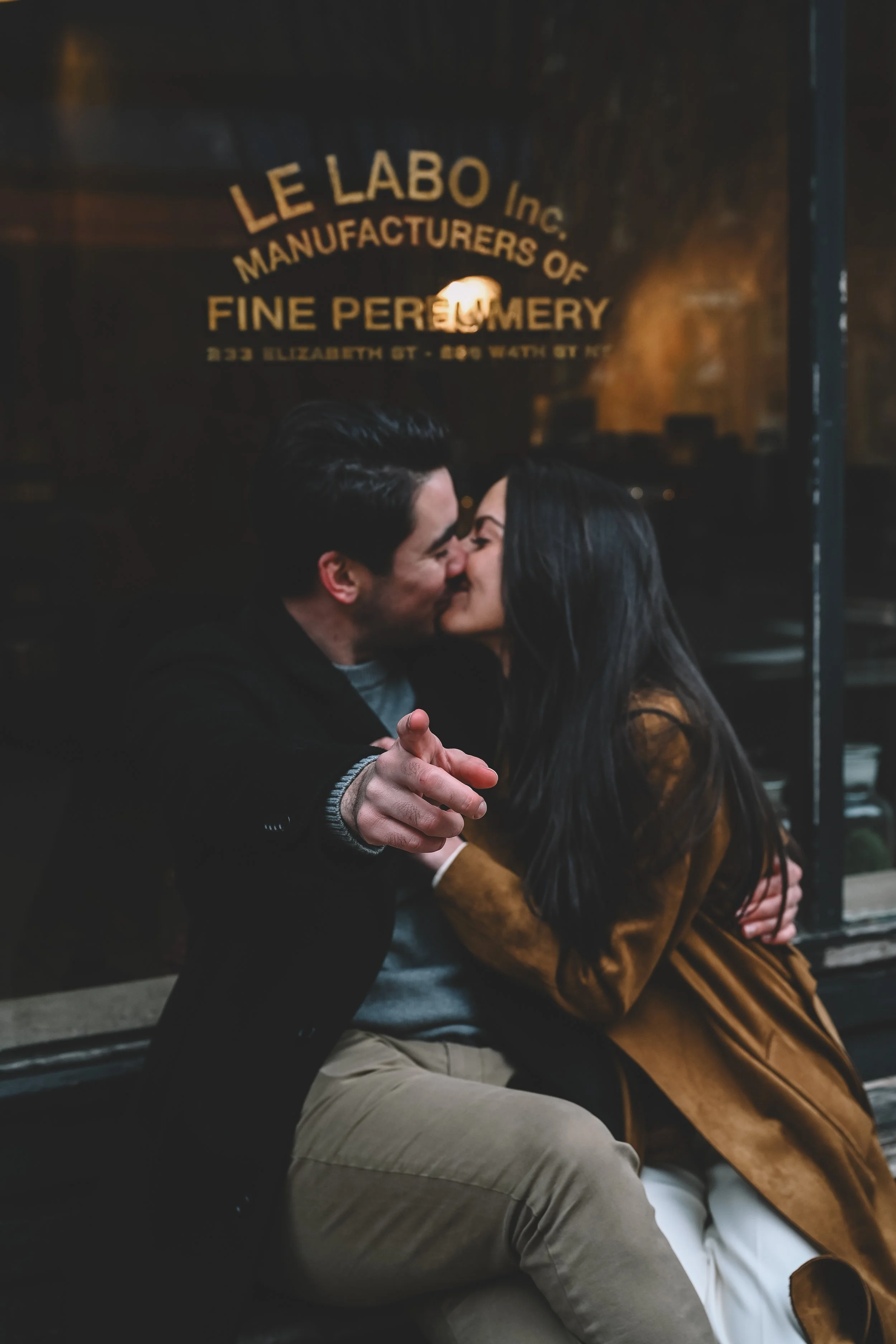 West Village engagement photos featuring a couple embracing outside a neighborhood storefront at night, captured in an atmospheric, artful style by NYC wedding photographer Robert Carlo.