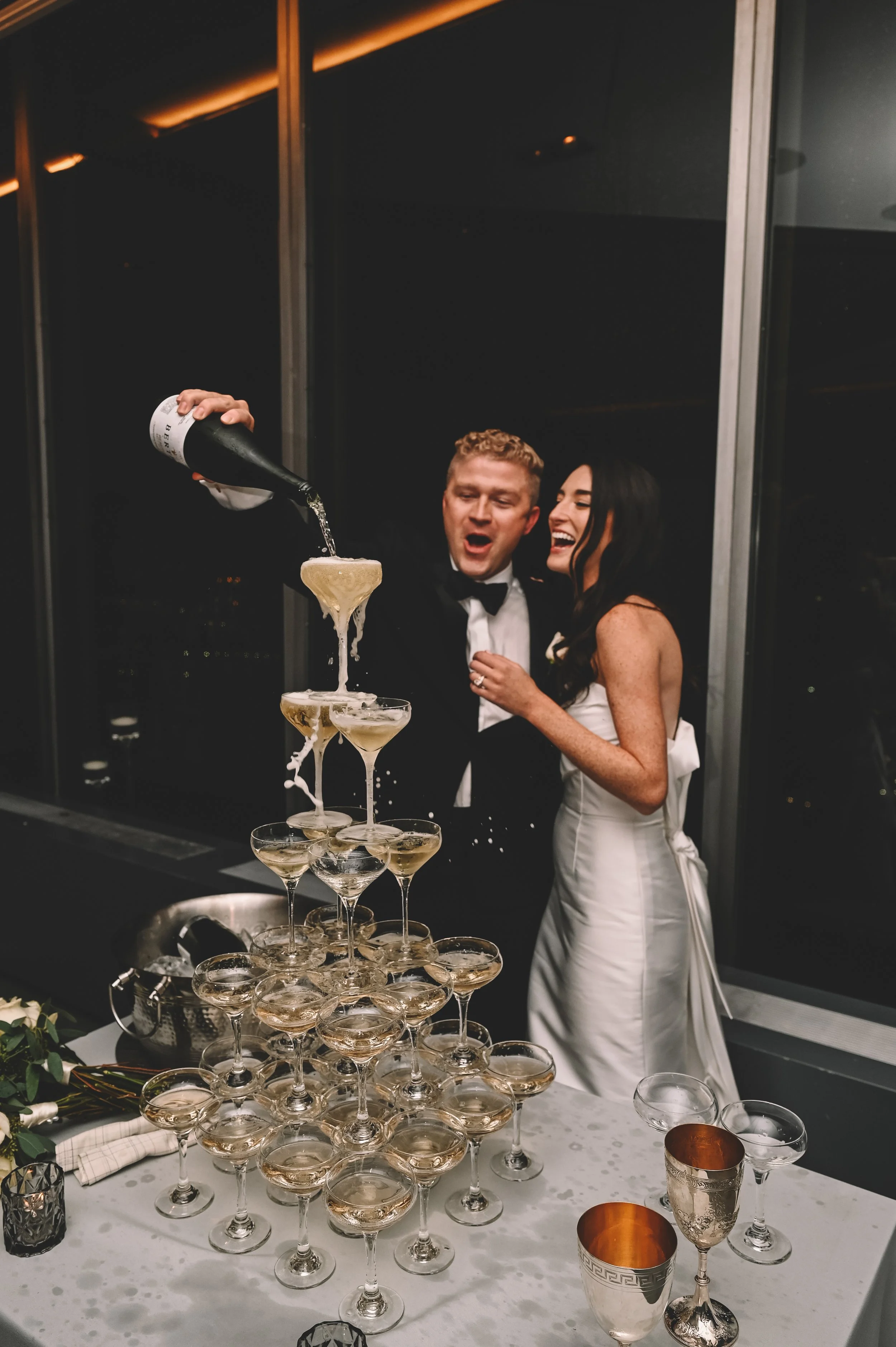 A bride and groom celebrating their wedding by pouring champagne into a pyramid of glasses. The bride is wearing a white wedding dress, and the groom is in a tuxedo. They are indoors with large windows behind them, and the scene is lively and joyful.