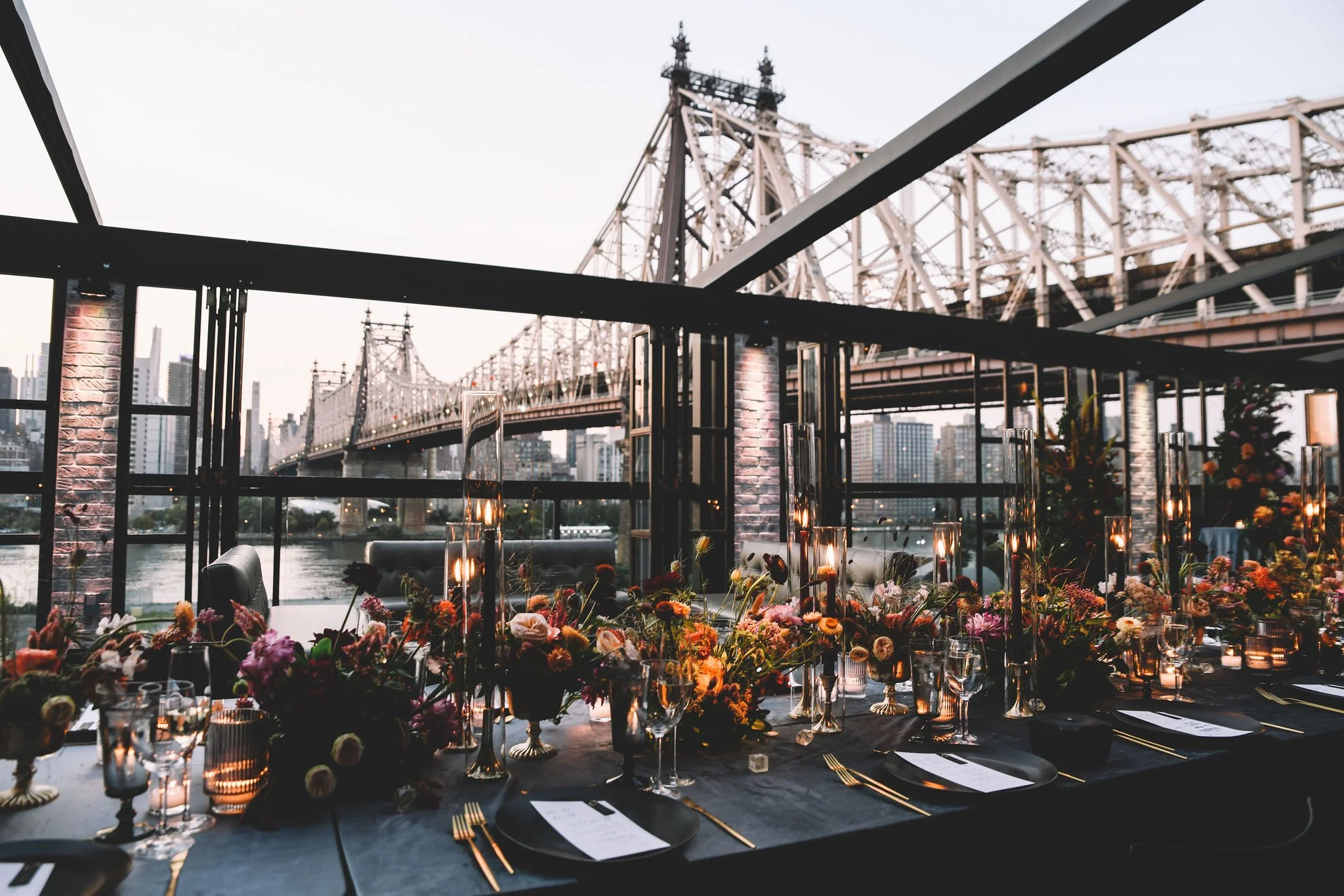 A wedding reception table decorated with flowers and candles, with a view of a bridge and city skyline through large windows.
