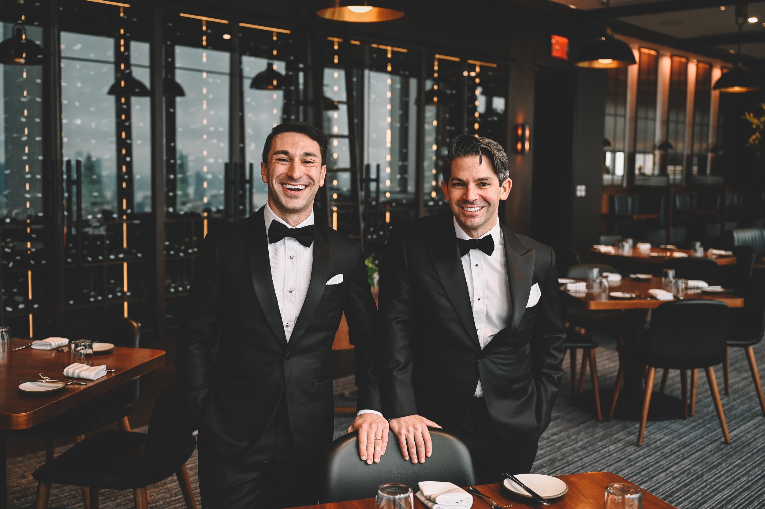 Two men in tuxedos smiling in a restaurant with large windows and modern decor.