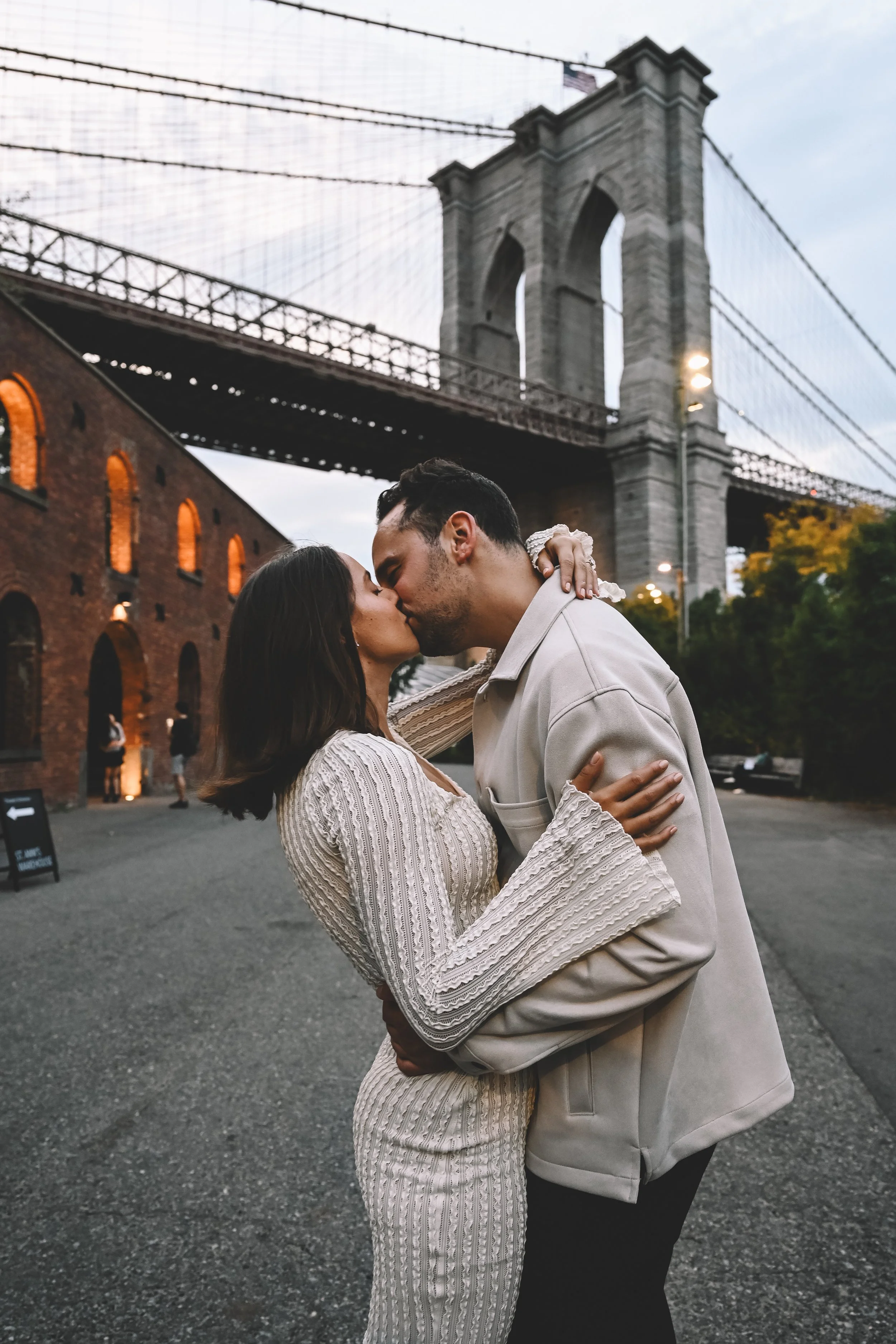 DUMBO engagement session featuring a couple kissing beneath the Brooklyn Bridge at dusk, captured in a timeless, atmospheric style by NYC engagement photographer Robert Carlo.