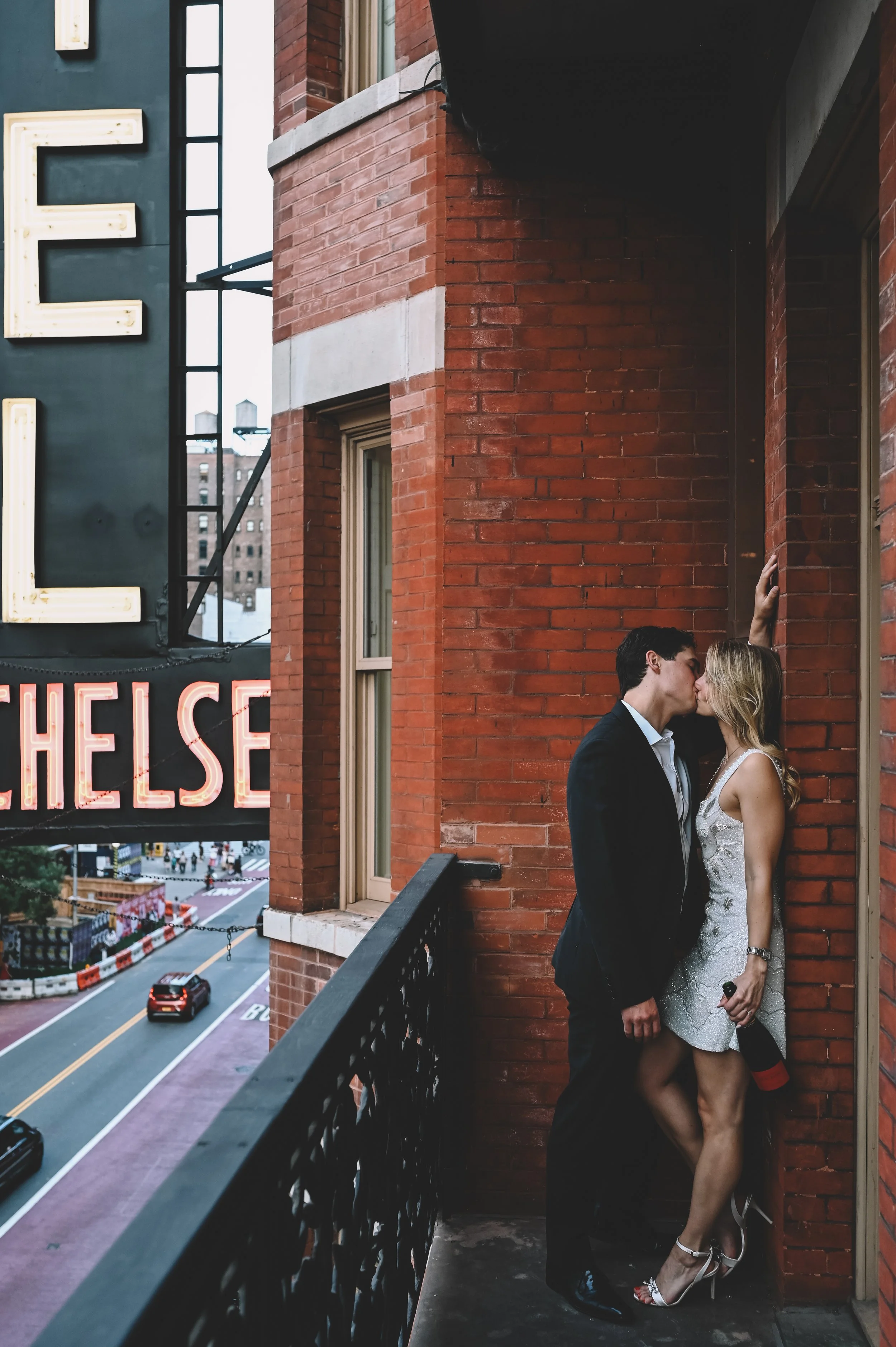 A couple kissing on a balcony against a red brick wall, with a city street and a neon sign that says 'CHELSEA' visible in the background.