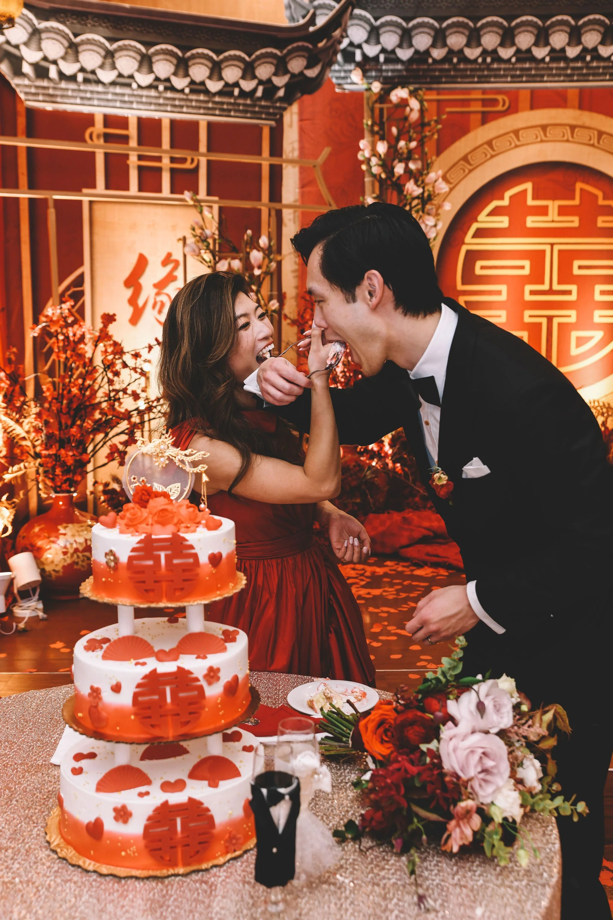 A couple in formal attire sharing a kiss during a celebration, with a three-tiered wedding cake decorated with red Chinese symbols and flowers, in an ornately decorated Asian-themed setting with red and gold accents and floral arrangements.