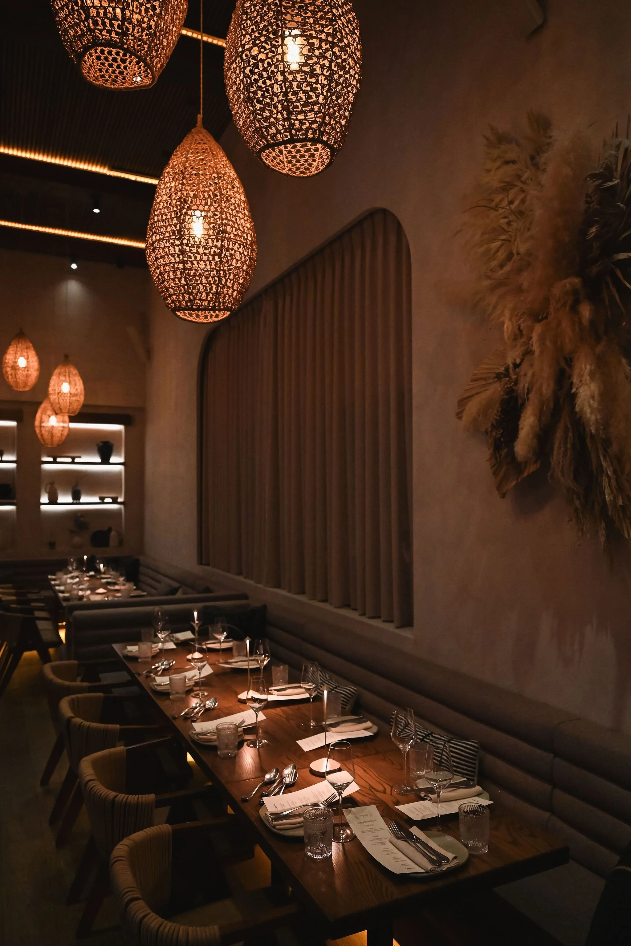 Elegant restaurant interior with hanging woven pendant lights, a long dining table set with wine glasses, silverware, napkins, and menus, and decorative elements on the wall.