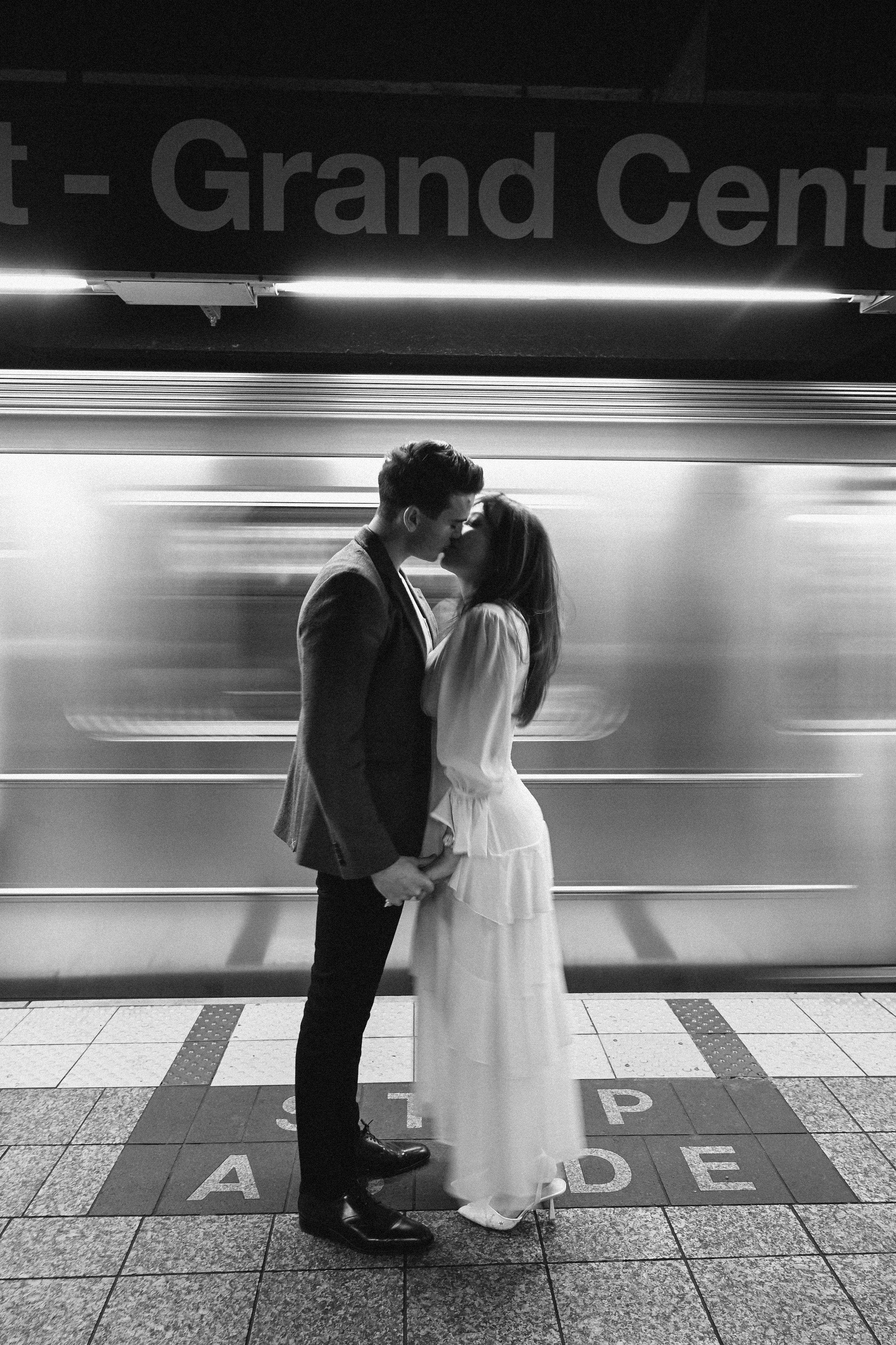 Black and white engagement photos at Grand Central Station in NYC, featuring a couple embracing on the subway platform, captured by NYC wedding photographer Robert Carlo.