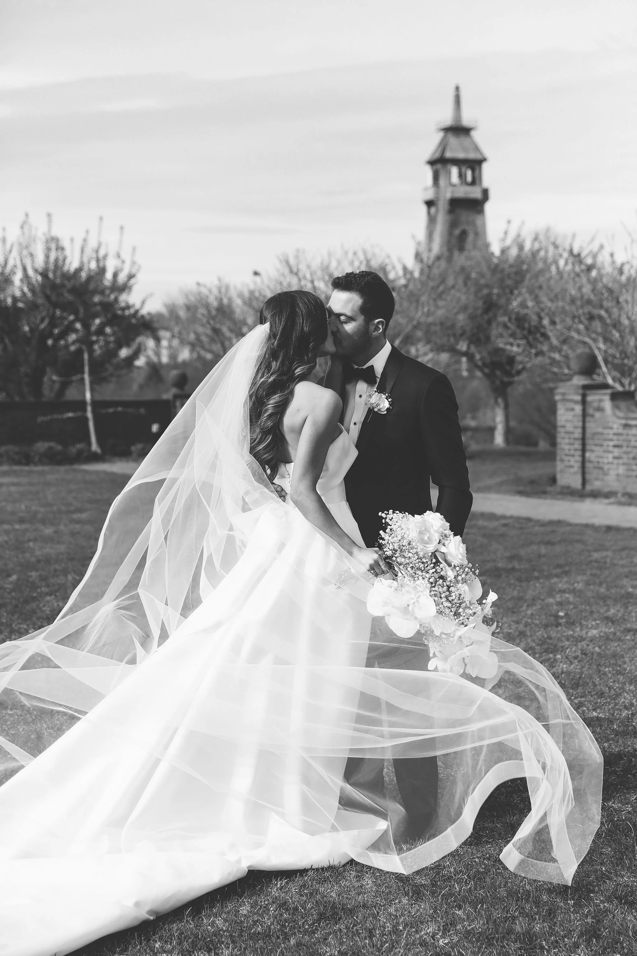 Black and white wedding photo of a bride and groom kissing outdoors, with the bride holding a bouquet of flowers and wearing a flowing wedding dress, and the groom in a tuxedo, standing on a grassy area with trees and a tower in the background.