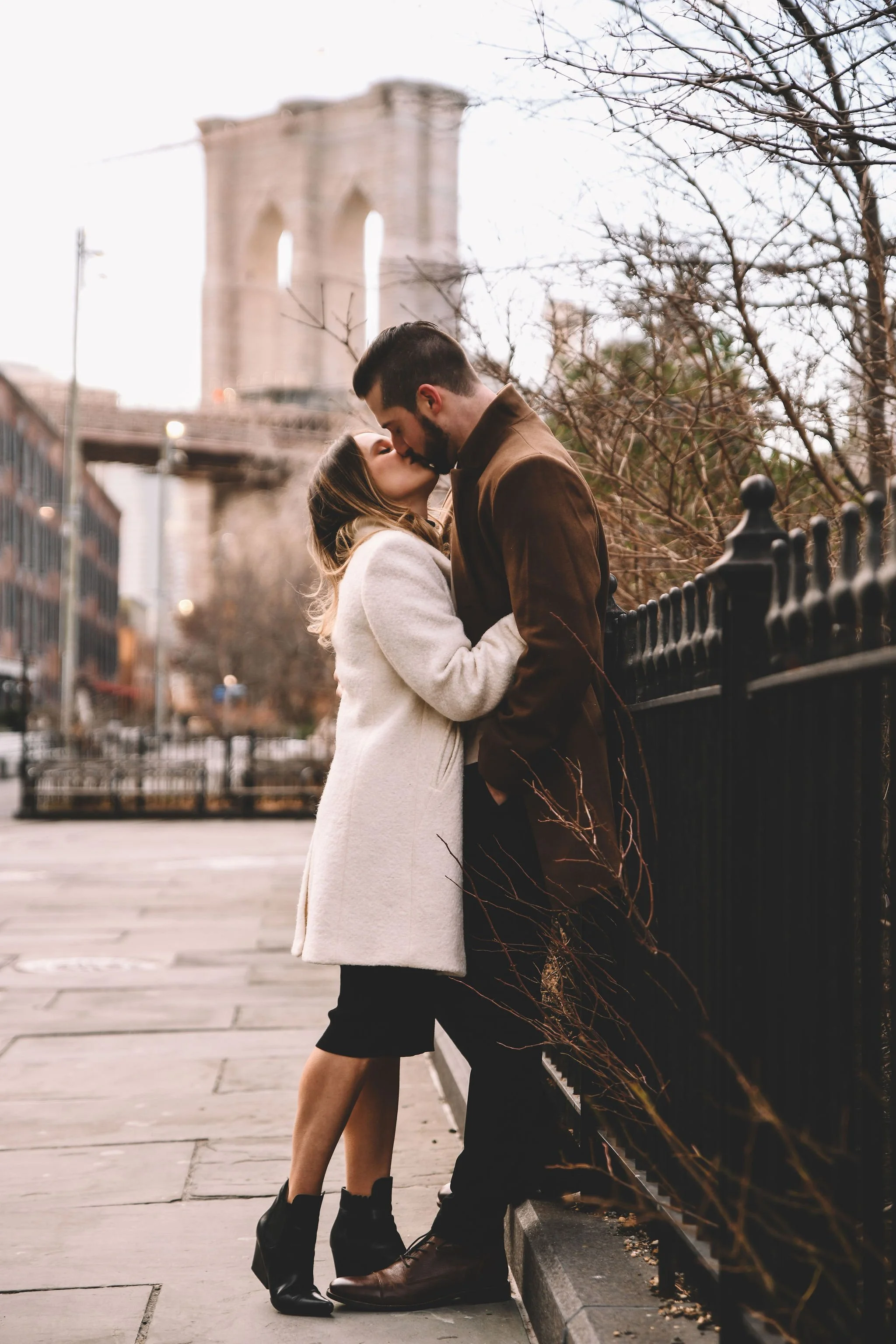 A couple is kissing on a city street, standing next to a black iron fence with leafless trees in the background and a large bridge structure behind them.