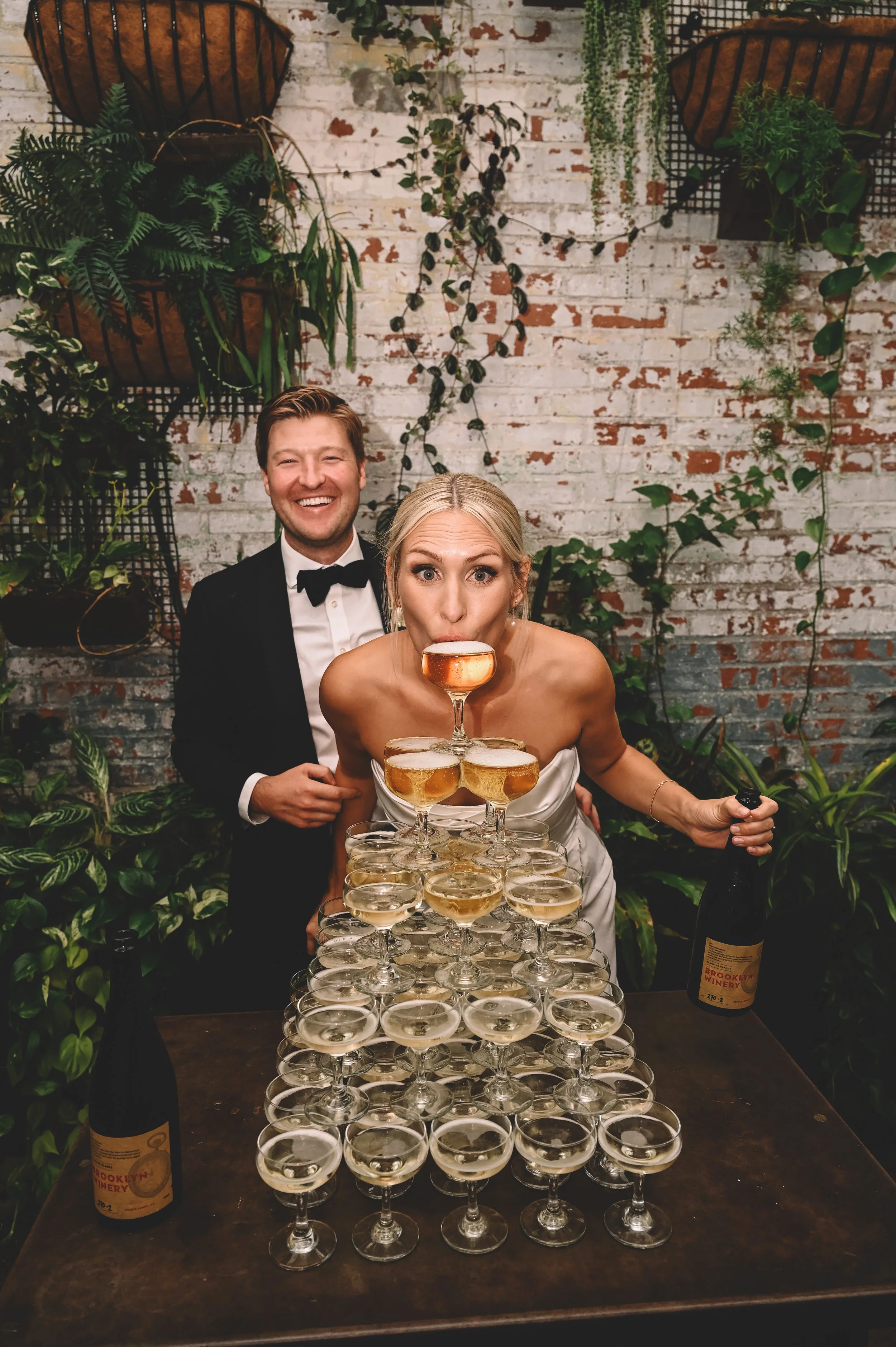 Bride sipping from the top glass of a champagne tower while the groom laughs behind her during a lively wedding reception at Brooklyn Winery in Williamsburg, captured in a candid, documentary style by NYC wedding photographer Robert Carlo.