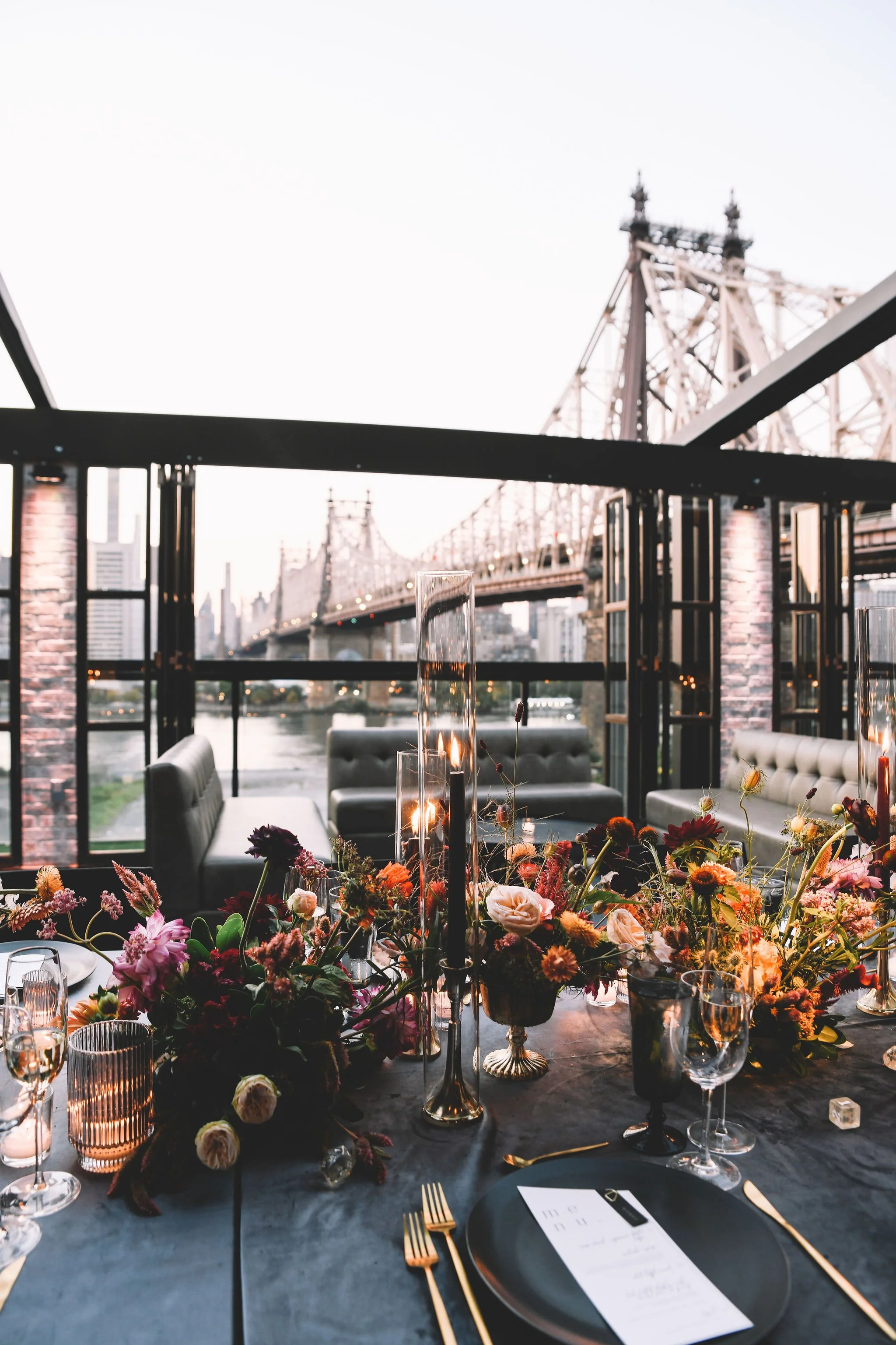 Elegant dining table set with flowers and candles, overlooking a bridge over a river at sunset.