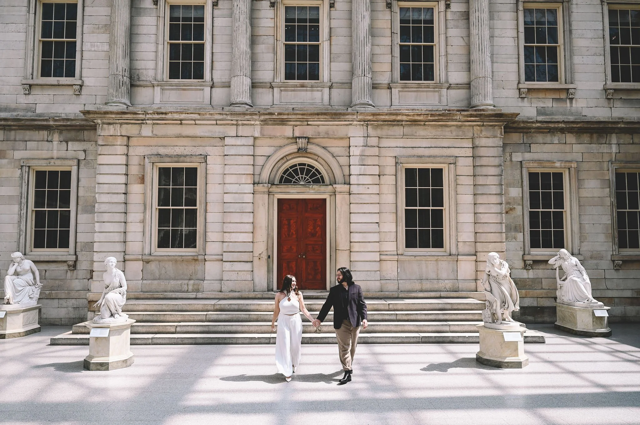 A couple holding hands walking in front of a historic building with classical architecture, including statues and columns.