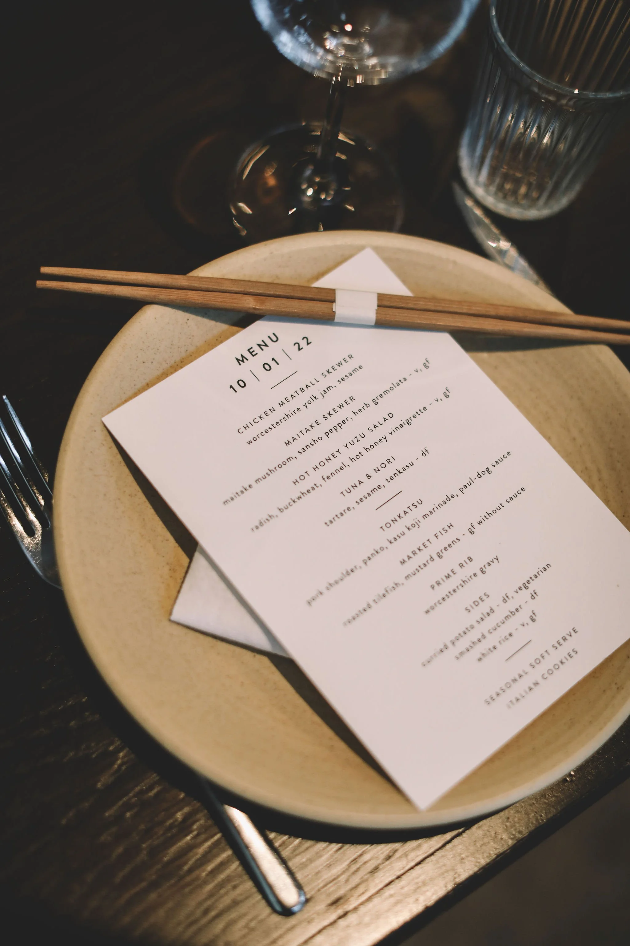 A restaurant table setting with a beige plate holding a menu and chopsticks, a wine glass, and a clear drinking glass, on a dark wooden surface.
