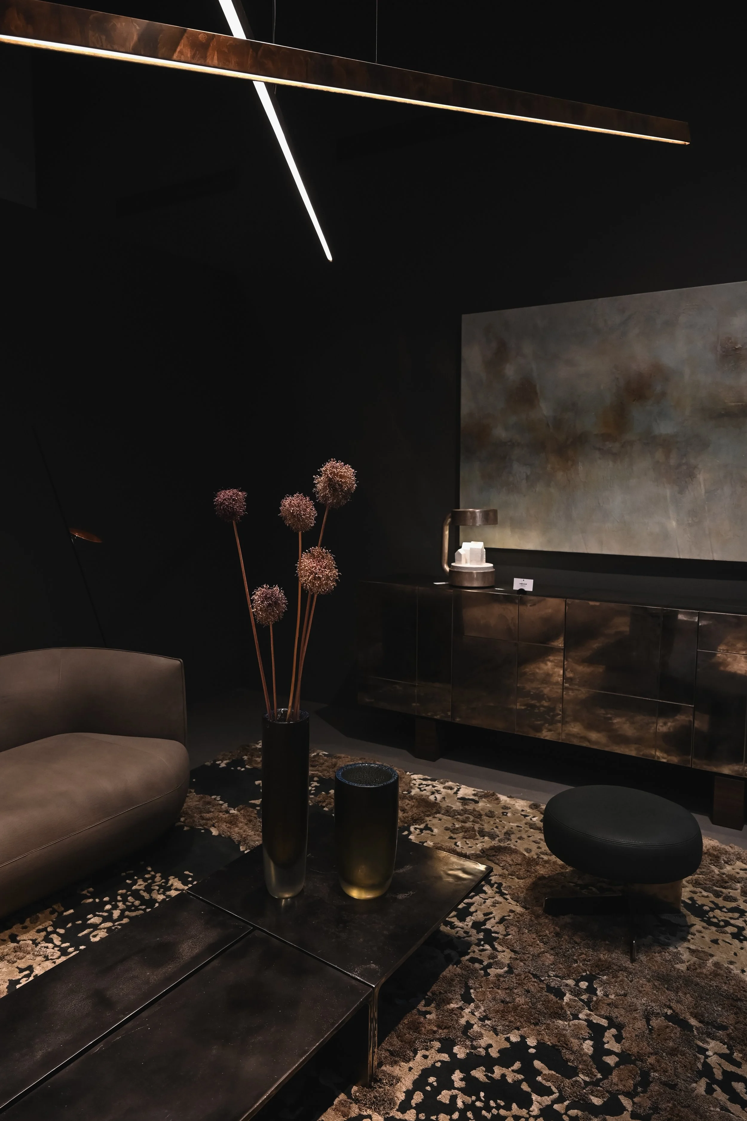 A modern, dark-colored living room with a leather sofa, a black metal coffee table with decorative vases, a patterned area rug, and a large abstract painting on the wall. There is a black cabinet with a small lamp and a minimalist ceiling light fixture.
