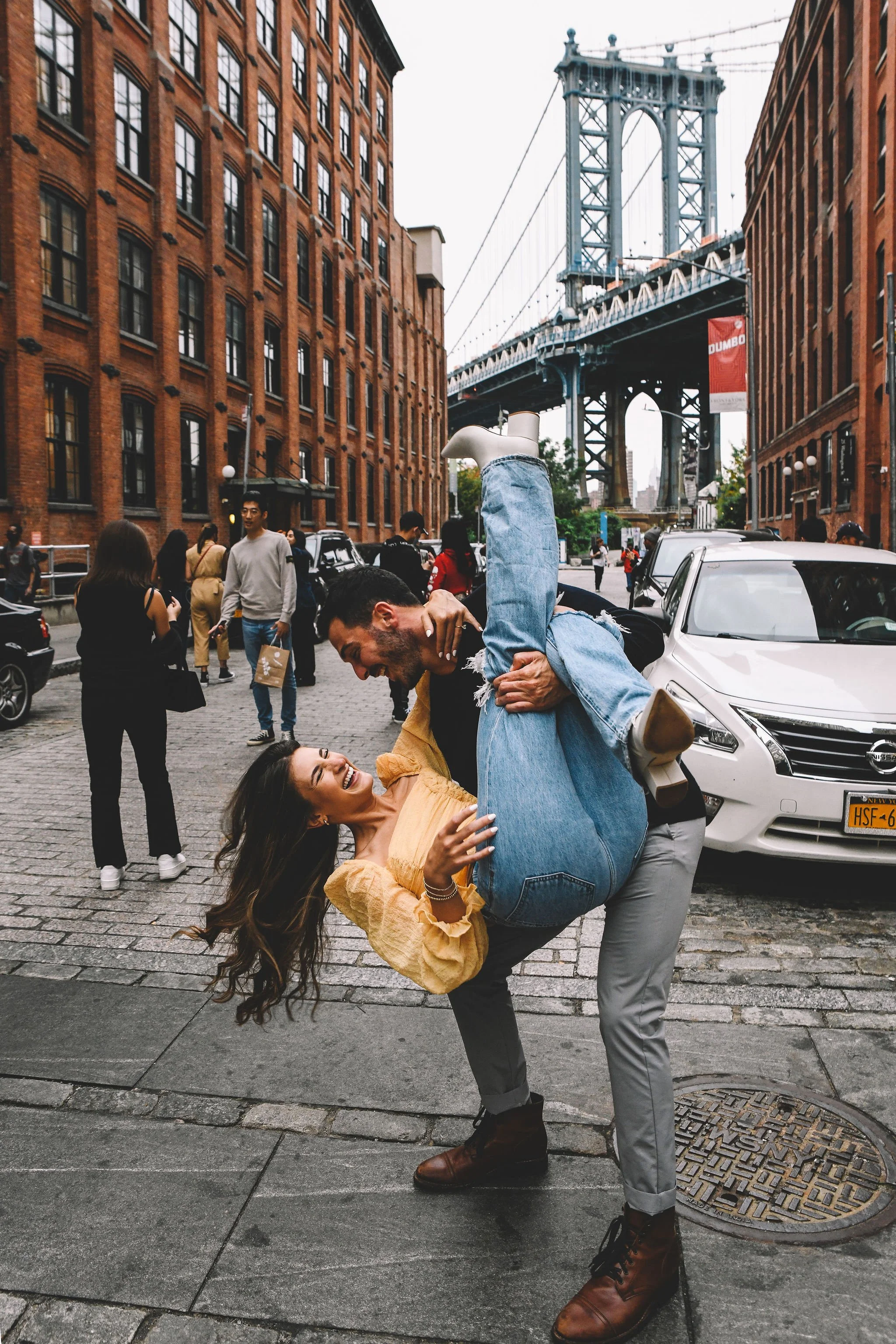 A couple dancing playfully on a city street with the Manhattan Bridge in the background, surrounded by pedestrians, parked cars, brick buildings, and an overcast sky.