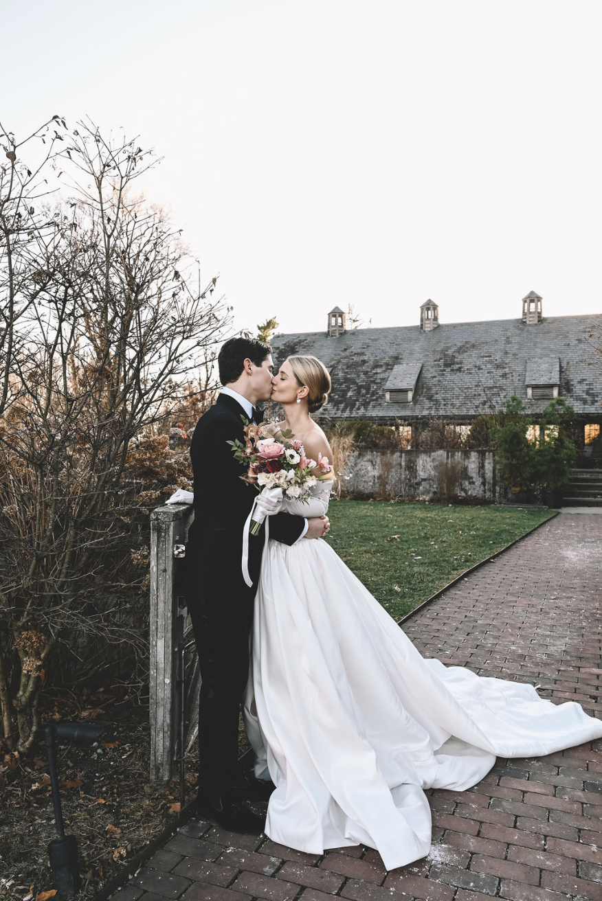 A bride and groom share a kiss outdoors on their wedding day, with the bride holding a bouquet of flowers, standing on a brick pathway near a rustic building and trees.