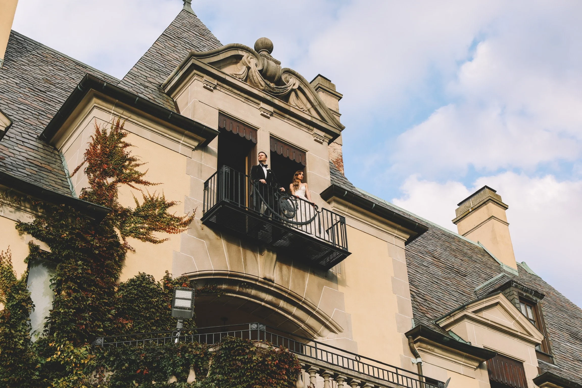 A couple dressed in wedding attire standing on a small balcony of a historic building with a slate roof and ivy growing on the wall, against a partly cloudy sky.