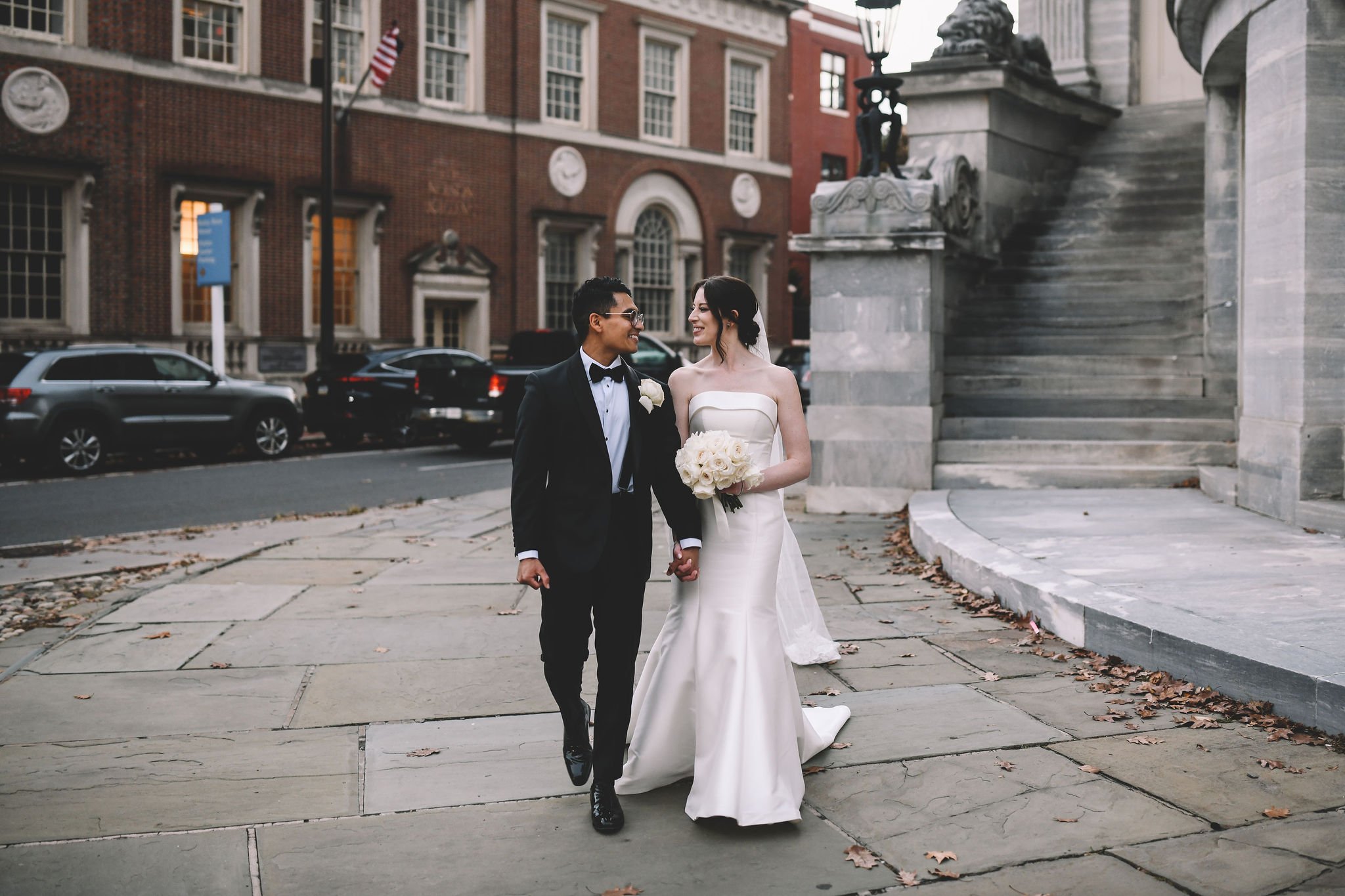 A newlywed couple walking hand in hand outside a courthouse, smiling at each other. The bride is in a white wedding gown holding a bouquet of white roses, and the groom is in a black tuxedo with a bow tie.