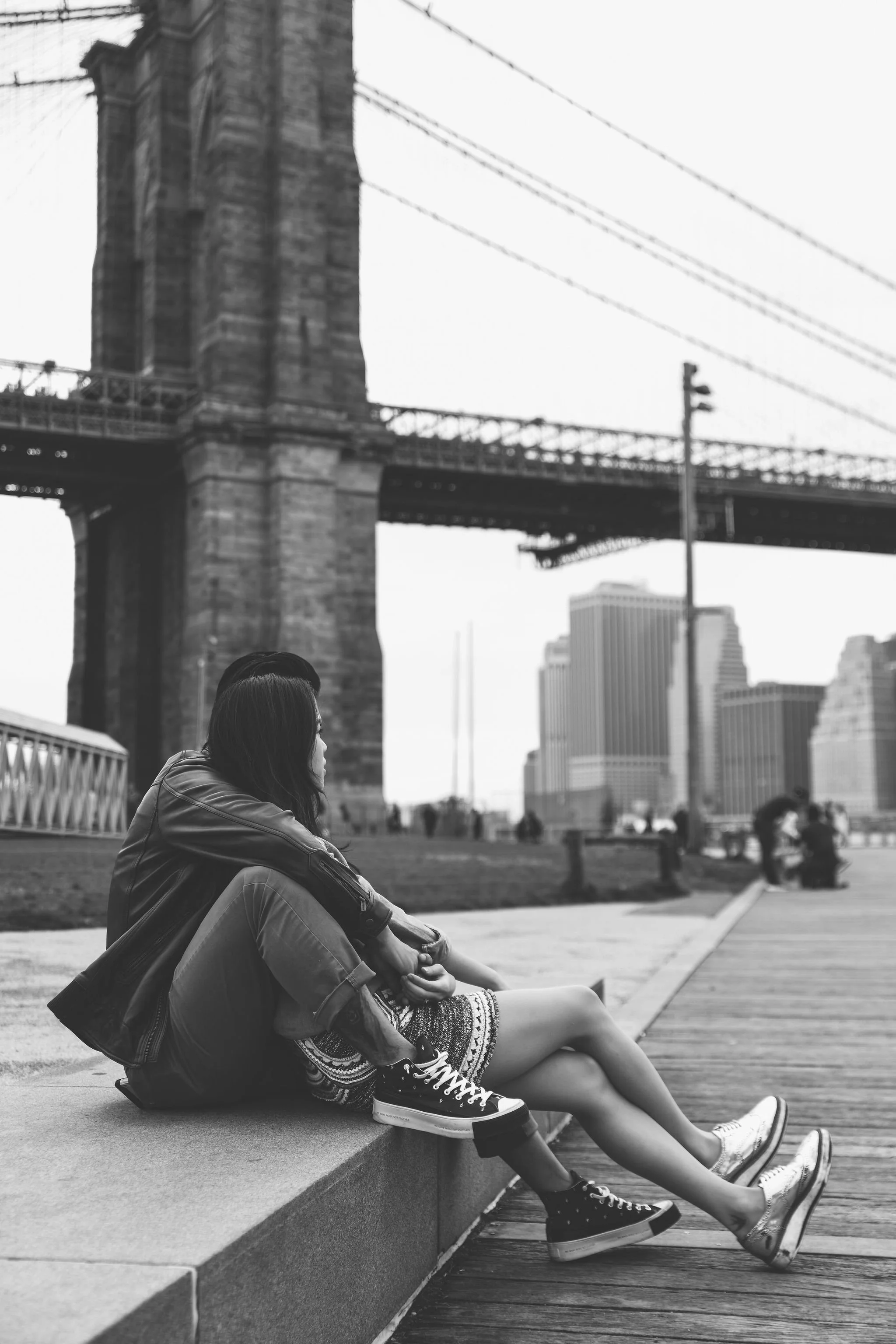 A black and white photo of two people sitting on a ledge near a body of water, with the Brooklyn Bridge and city buildings in the background.