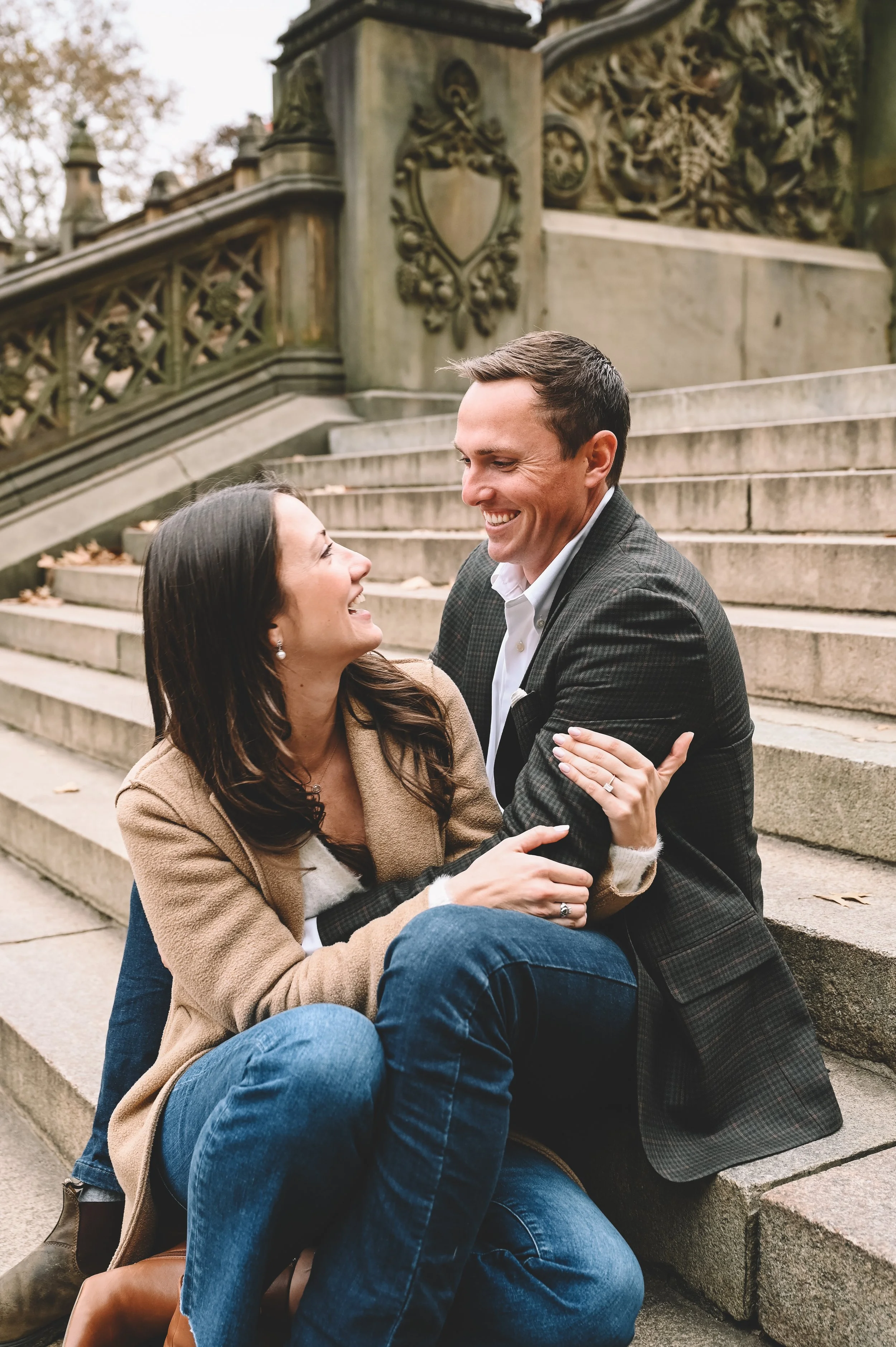 A couple smiling and holding each other on stone steps outdoors, with decorative stonework in the background.