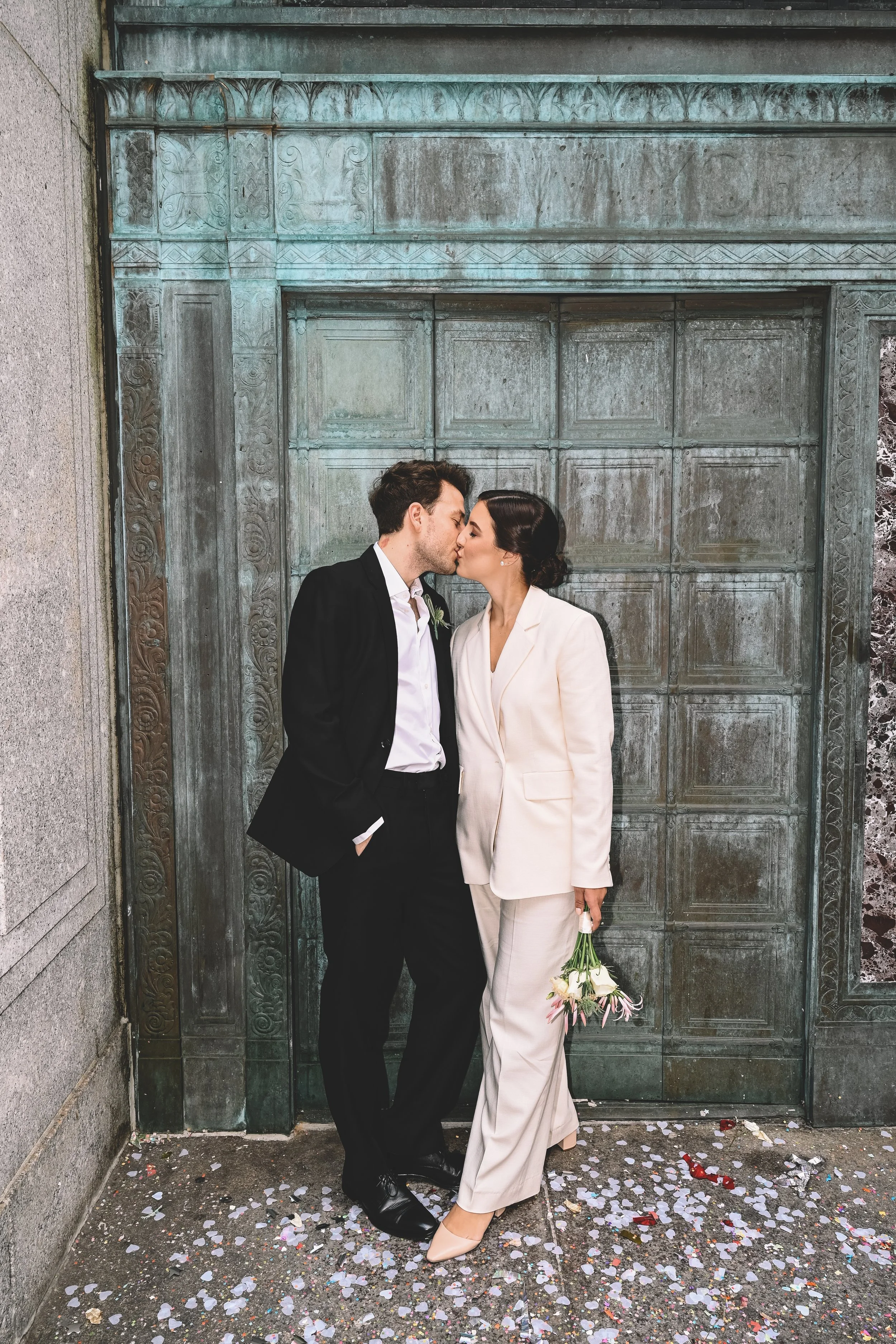 A couple dressed in formal attire sharing a kiss in front of a vintage green door, with confetti scattered on the ground, during a wedding celebration.